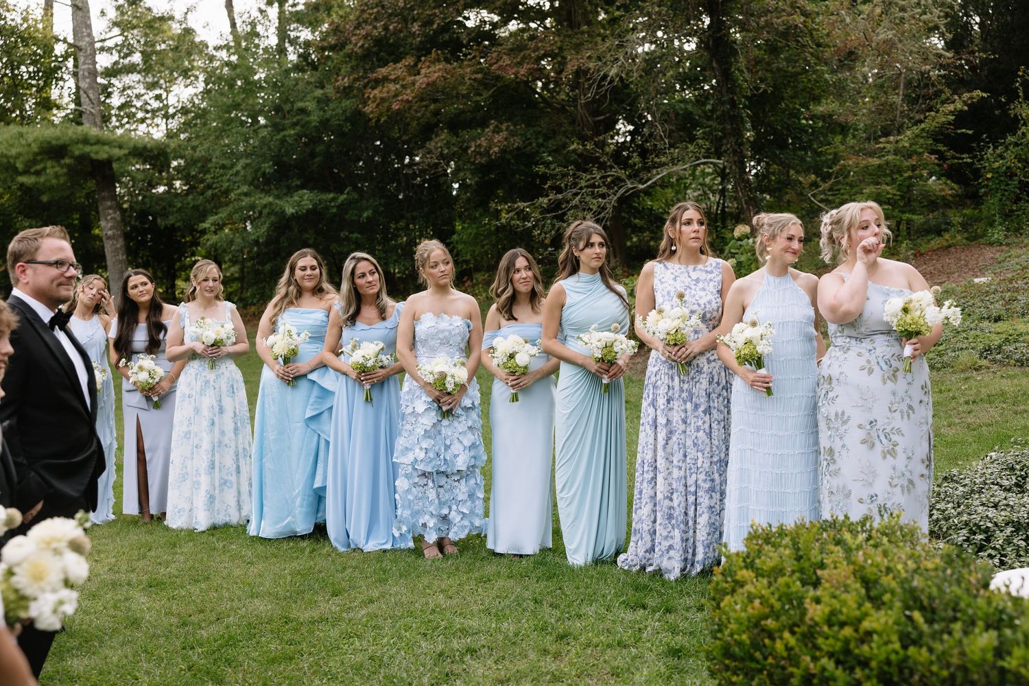 Bridal party in mixed blue and floral dresses poses on garden lawn.