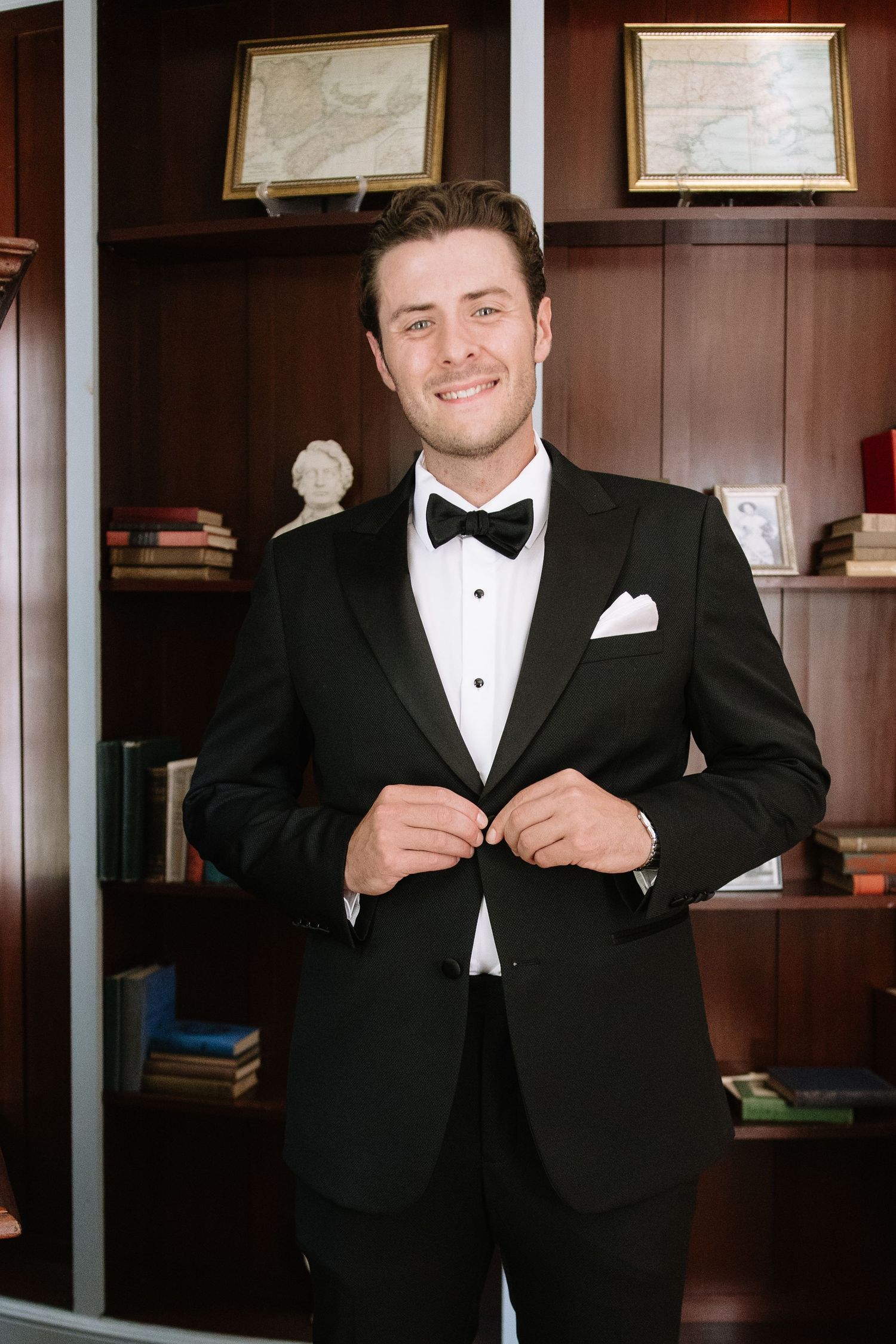 Groom in classic black tuxedo and bow tie smiles while adjusting his collar in a wood-paneled room.