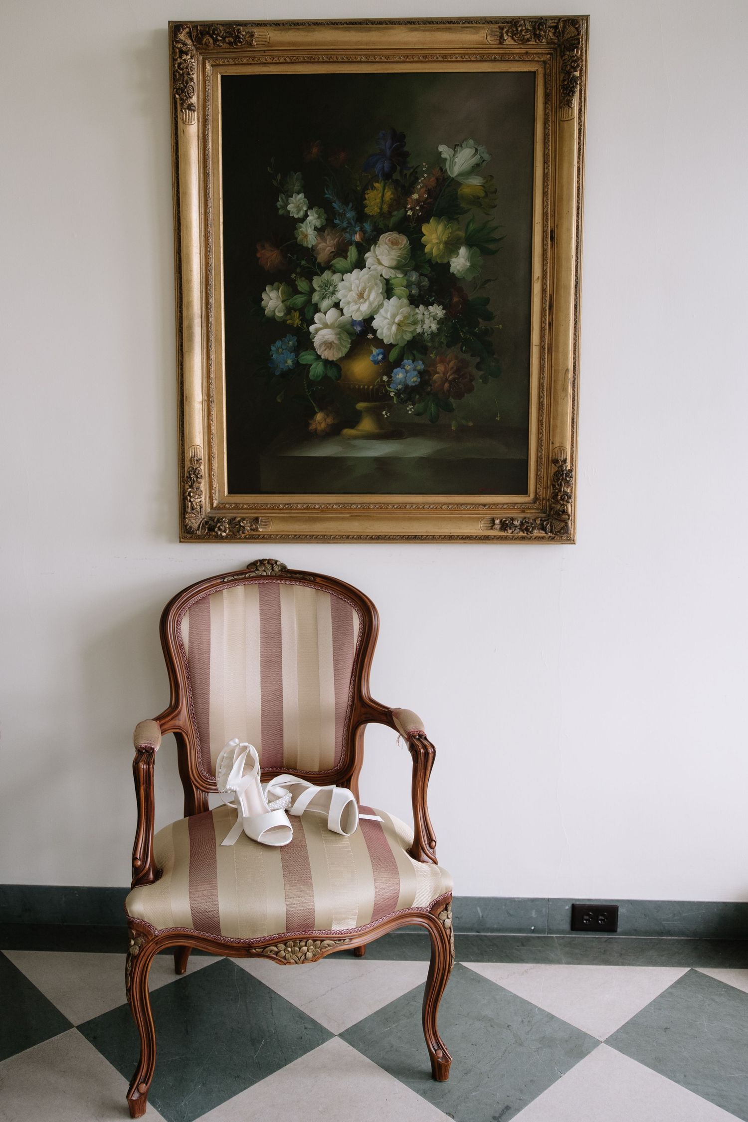Elegant striped chair sits beneath a classical floral painting in an ornate gold frame.