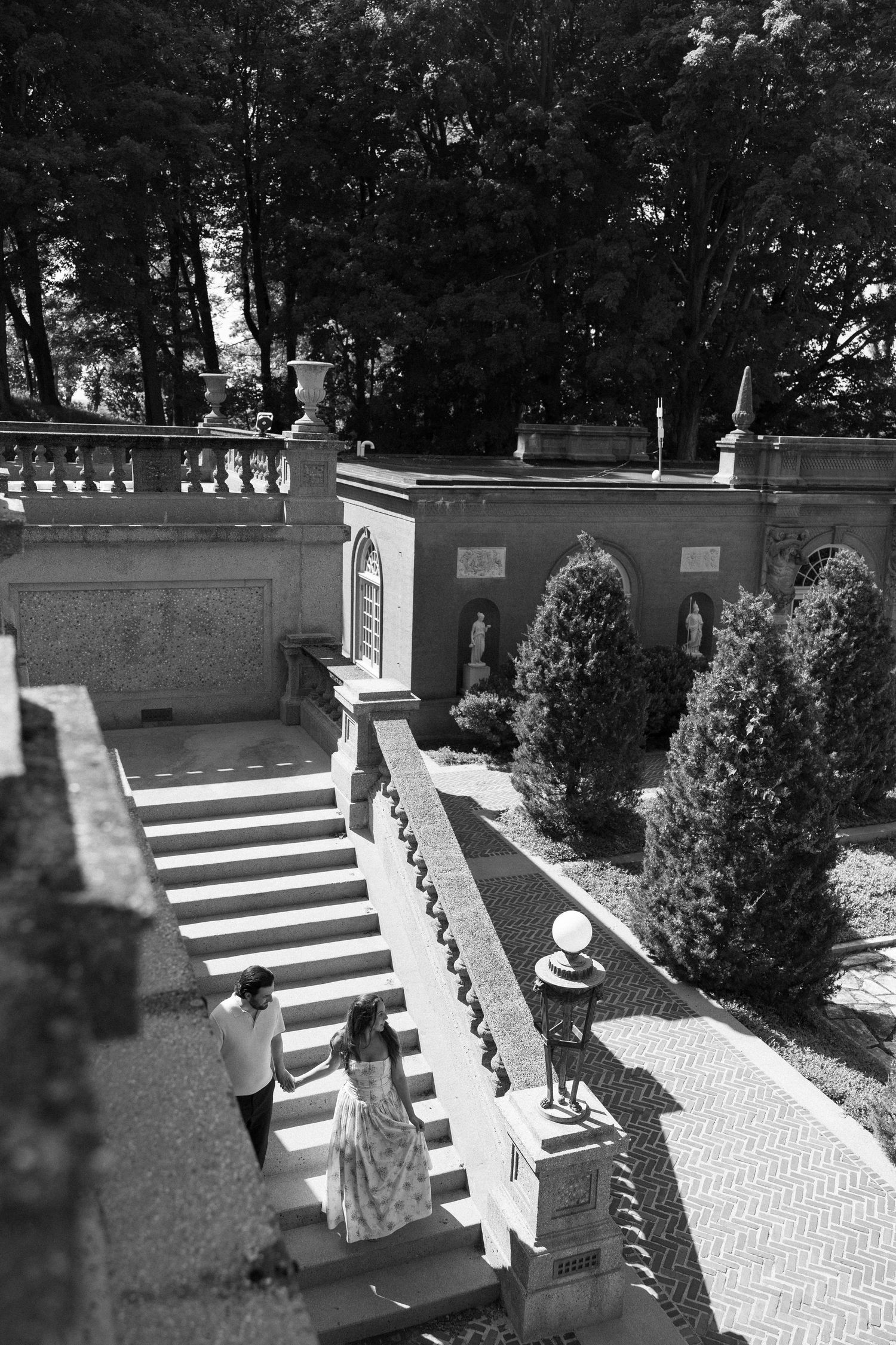 Couple ascending elegant stone staircase in formal garden with sculpted hedges and classical balustrade, black and white photo.