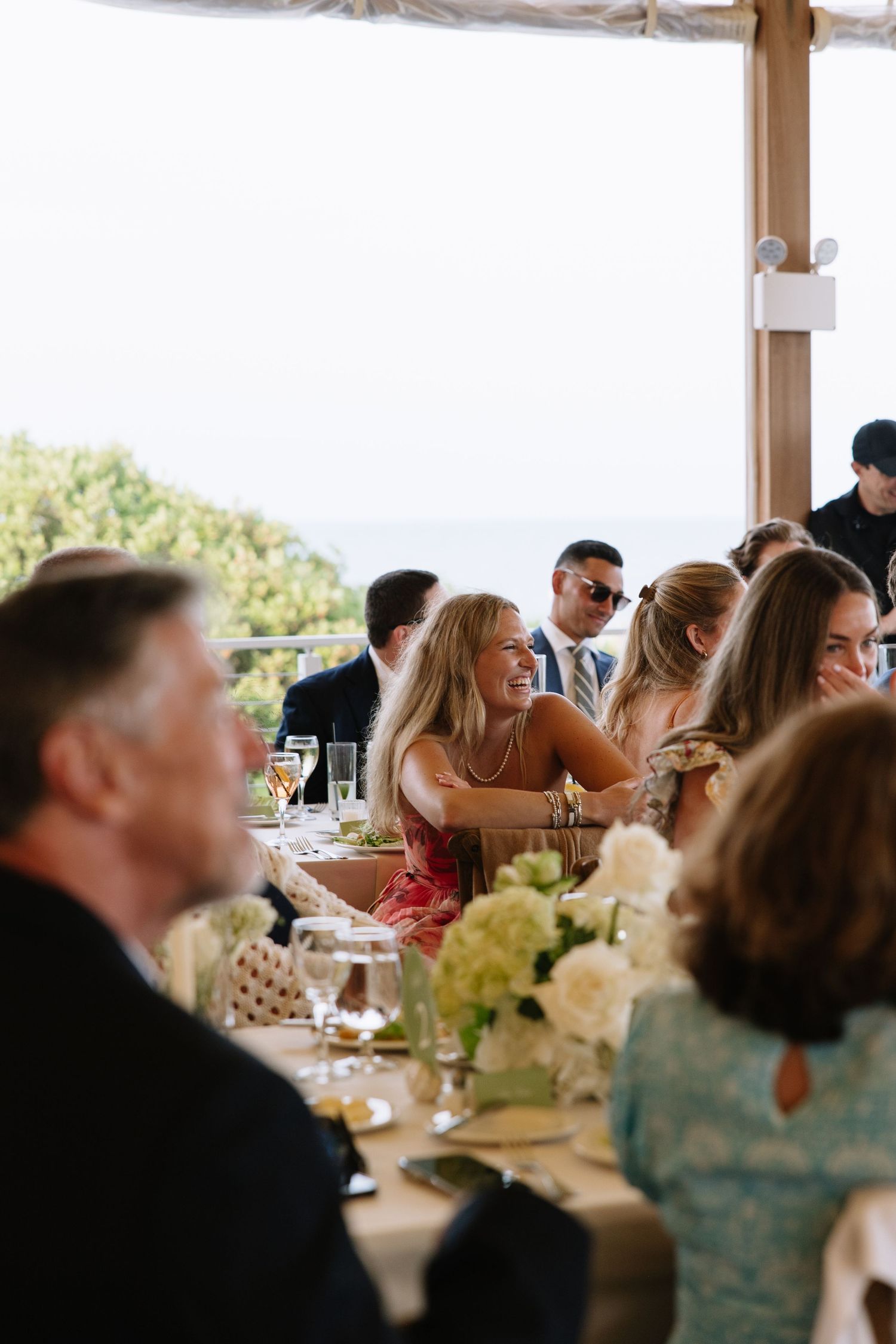 Guests laughing and socializing at an elegant outdoor wedding reception with ocean views and floral centerpieces.