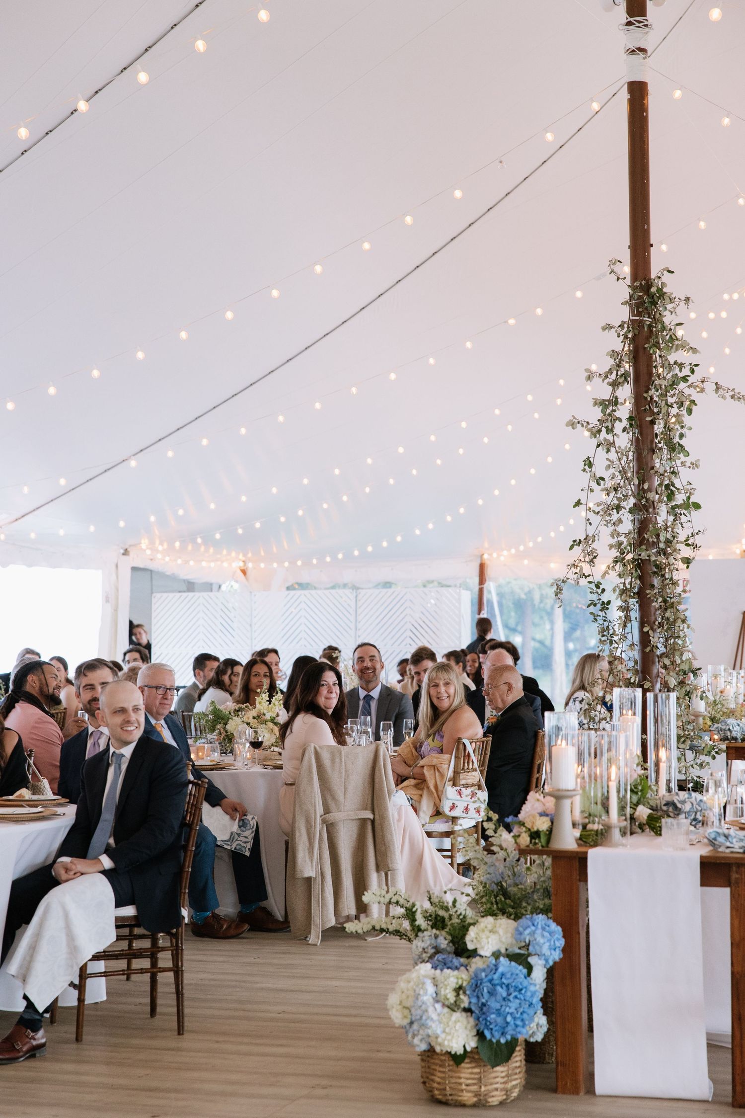 Elegant tented wedding reception with string lights, blue hydrangea centerpieces, and guests dining at long tables.