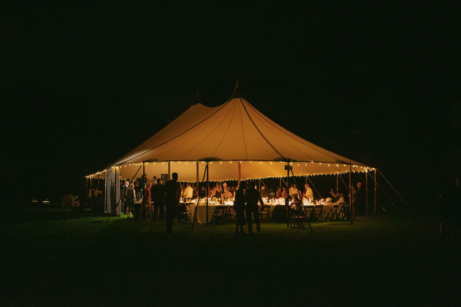 Illuminated wedding tent glowing warmly against a dark night sky with guests dining inside.
