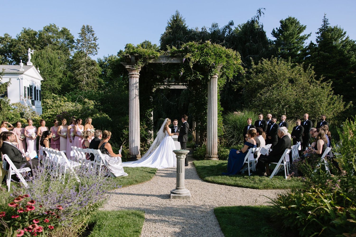 Elegant outdoor wedding ceremony under a vine-covered pergola with stone columns, surrounded by lush gardens and seated guests.