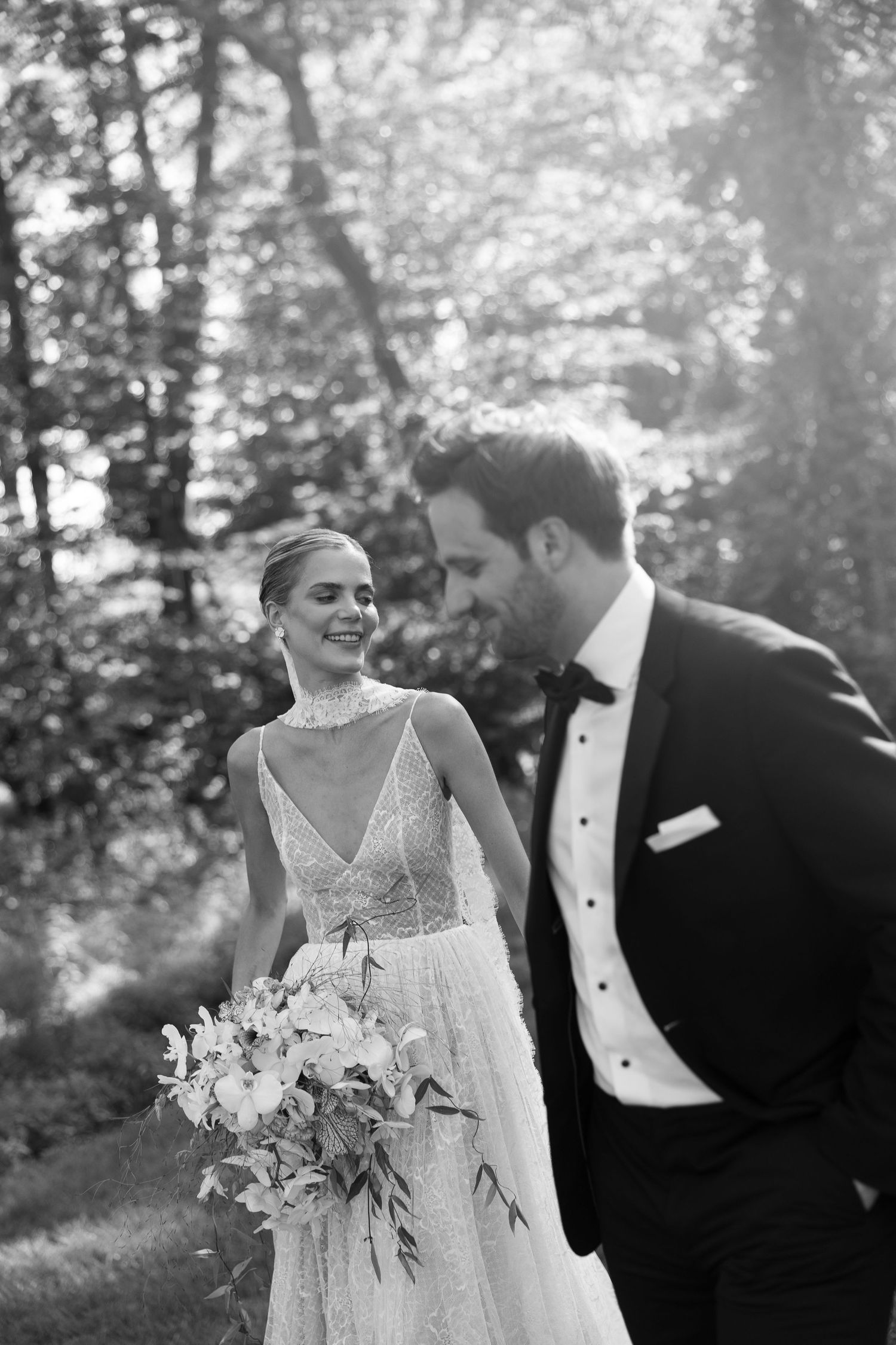 Black and white photo of a smiling bride in a lace gown holding a bouquet, walking with a groom in a tuxedo outdoors.