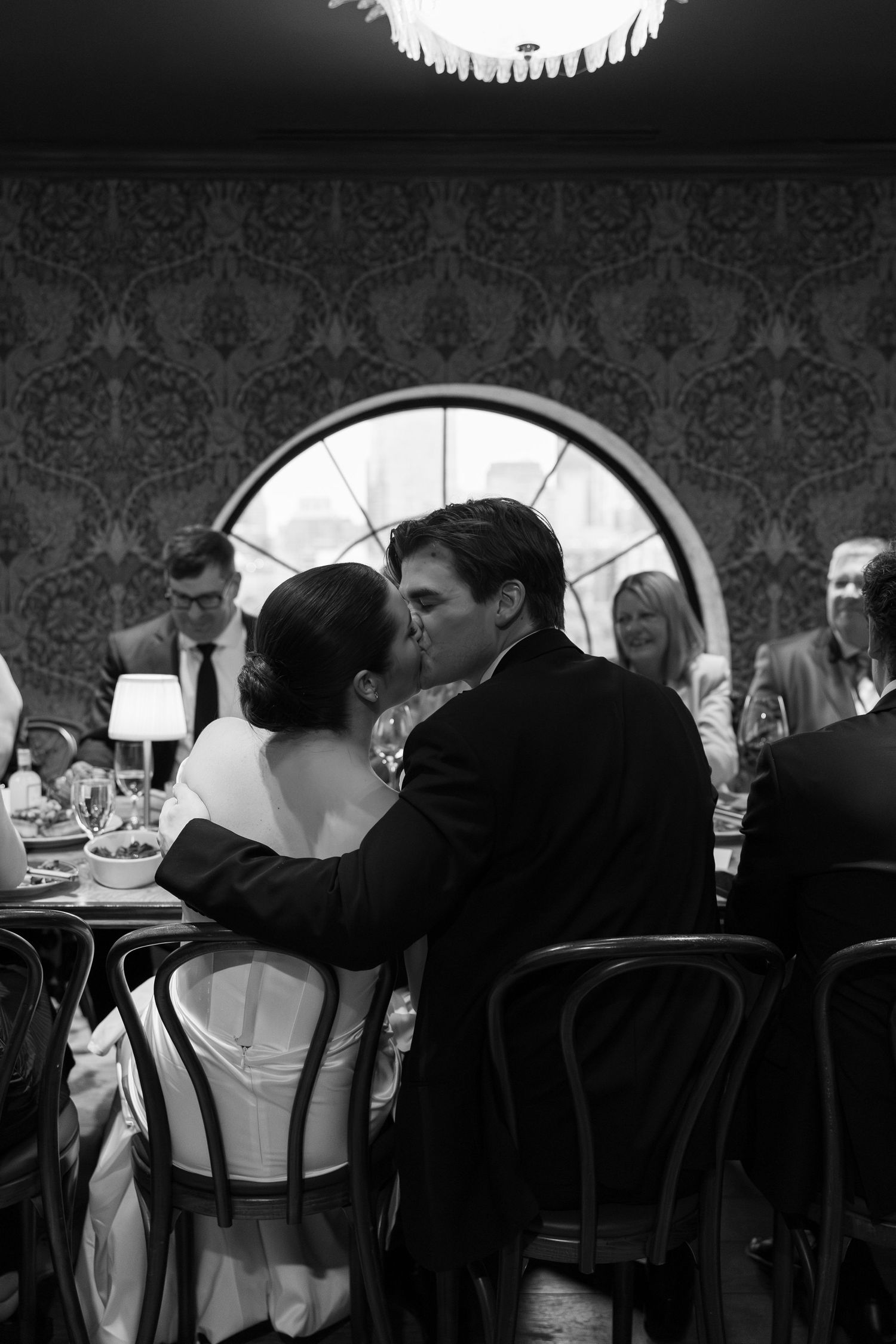 Black and white photo of newlyweds sharing a kiss at their wedding reception dinner table.