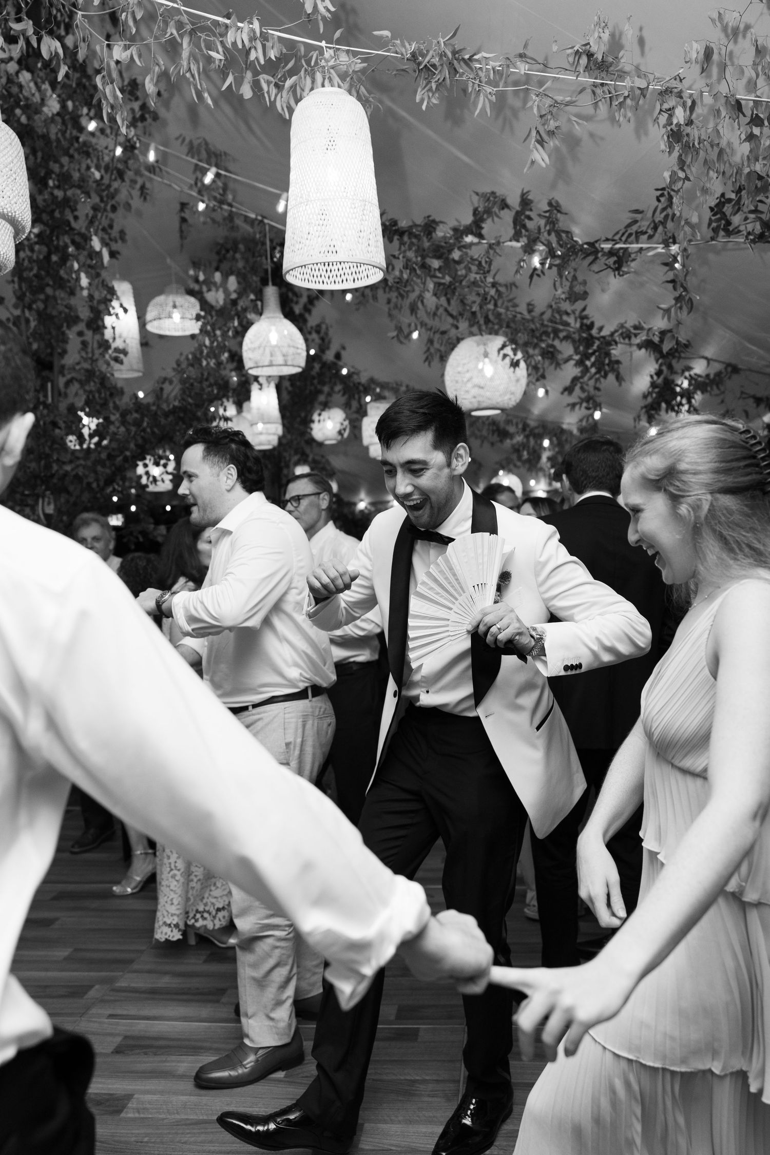 Black and white photo of groom in white tuxedo jacket dancing energetically with guests at wedding reception.