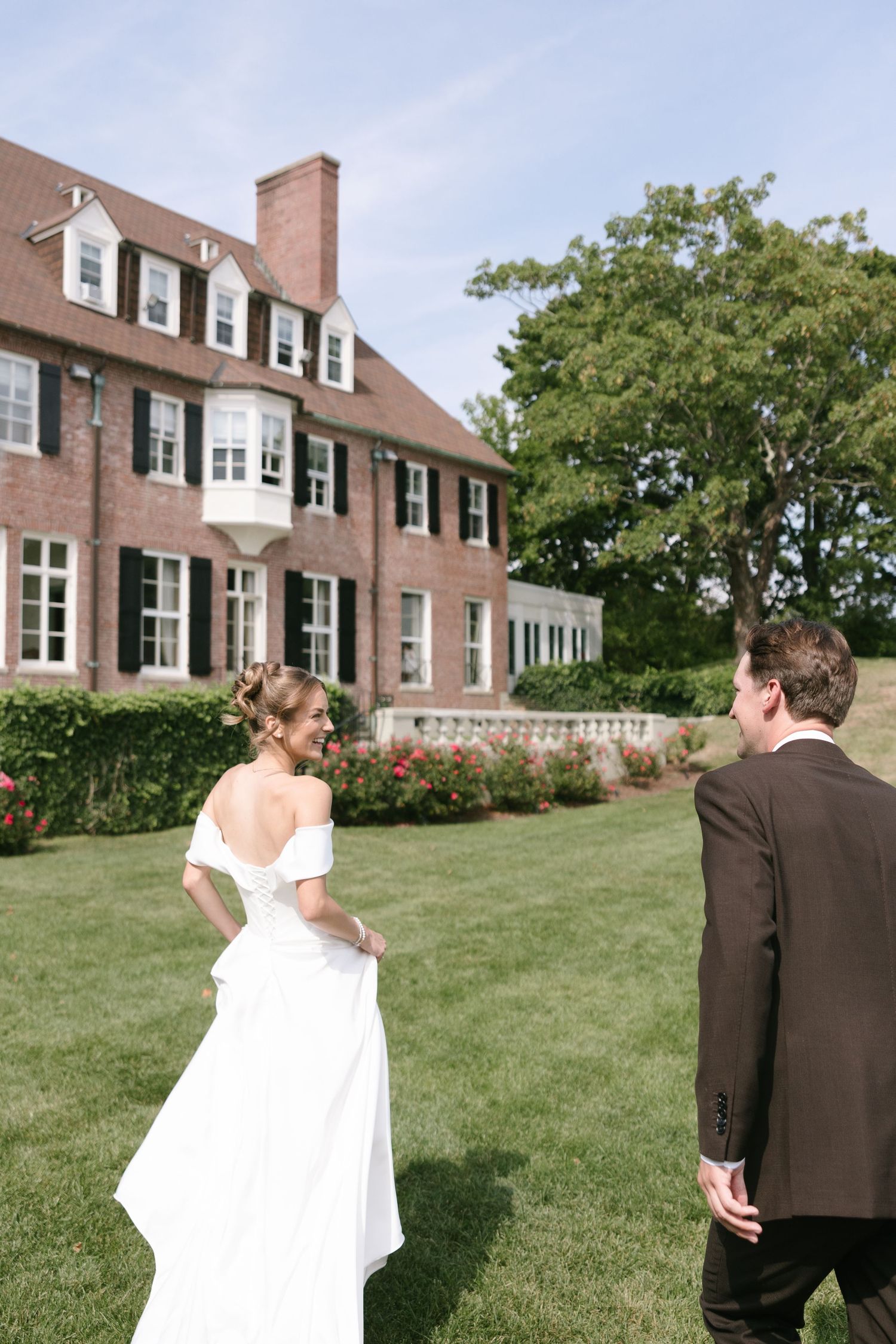 A bride in an off-the-shoulder white gown walks across a green lawn with a groom toward a historic brick mansion.