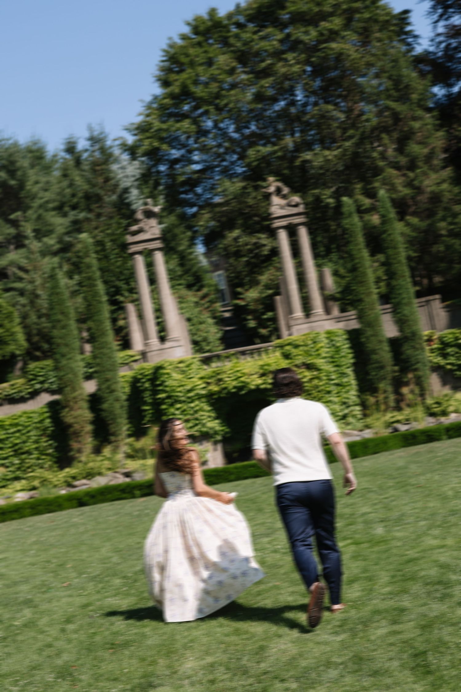 Bride in white gown and groom in white shirt run across lush green lawn in elegant garden with stone pillars.