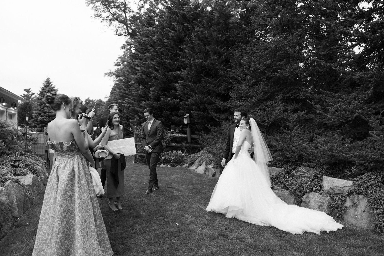 Black and white photo of wedding party gathering outdoors in a lush garden setting.