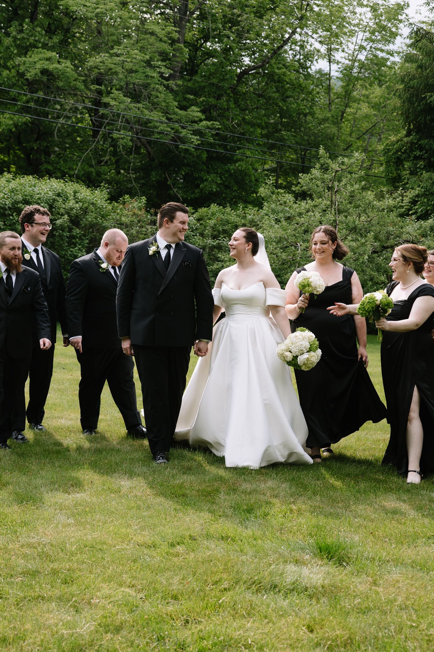 A bride and groom walk with their wedding party dressed in black on a green lawn surrounded by lush trees.