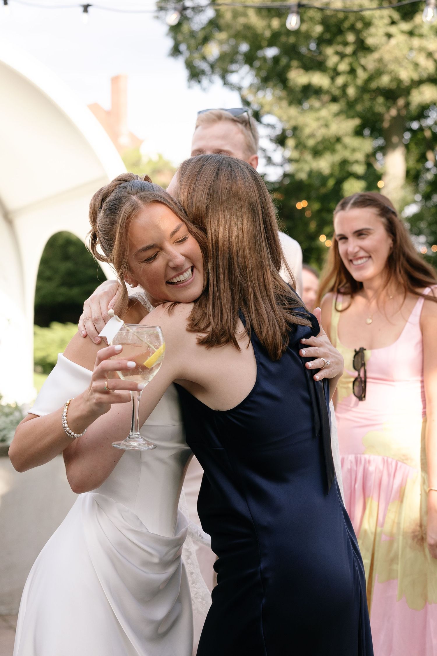 Laughing bride in a white dress hugs a guest in a navy dress while holding a drink at an outdoor wedding reception.