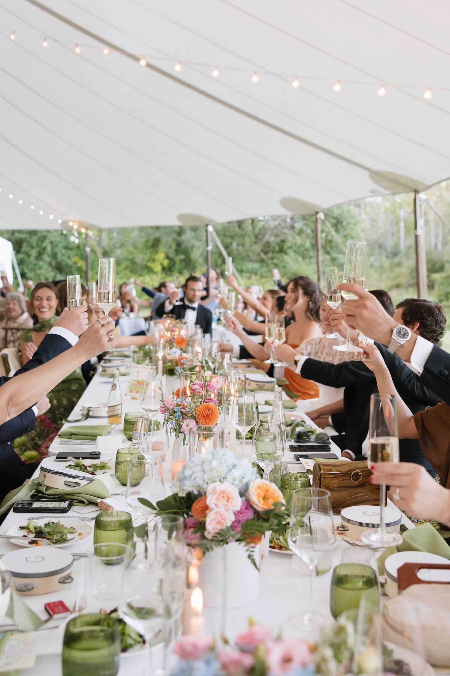 Wedding guests raise champagne glasses in a toast at a long decorated table under a white tent.