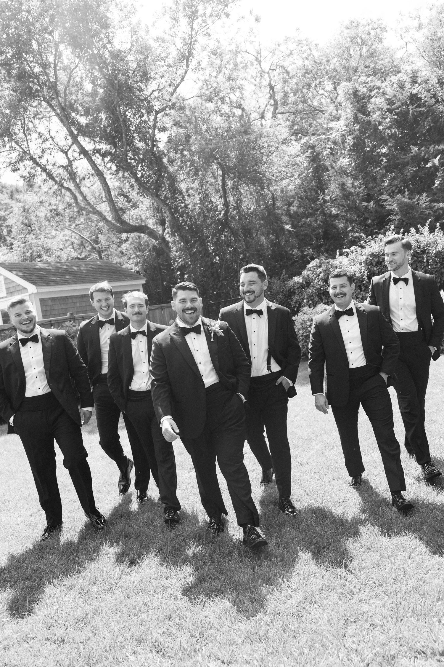 Seven groomsmen in black tuxedos and bow ties walk together laughing on a lawn in a classic black and white wedding photo.