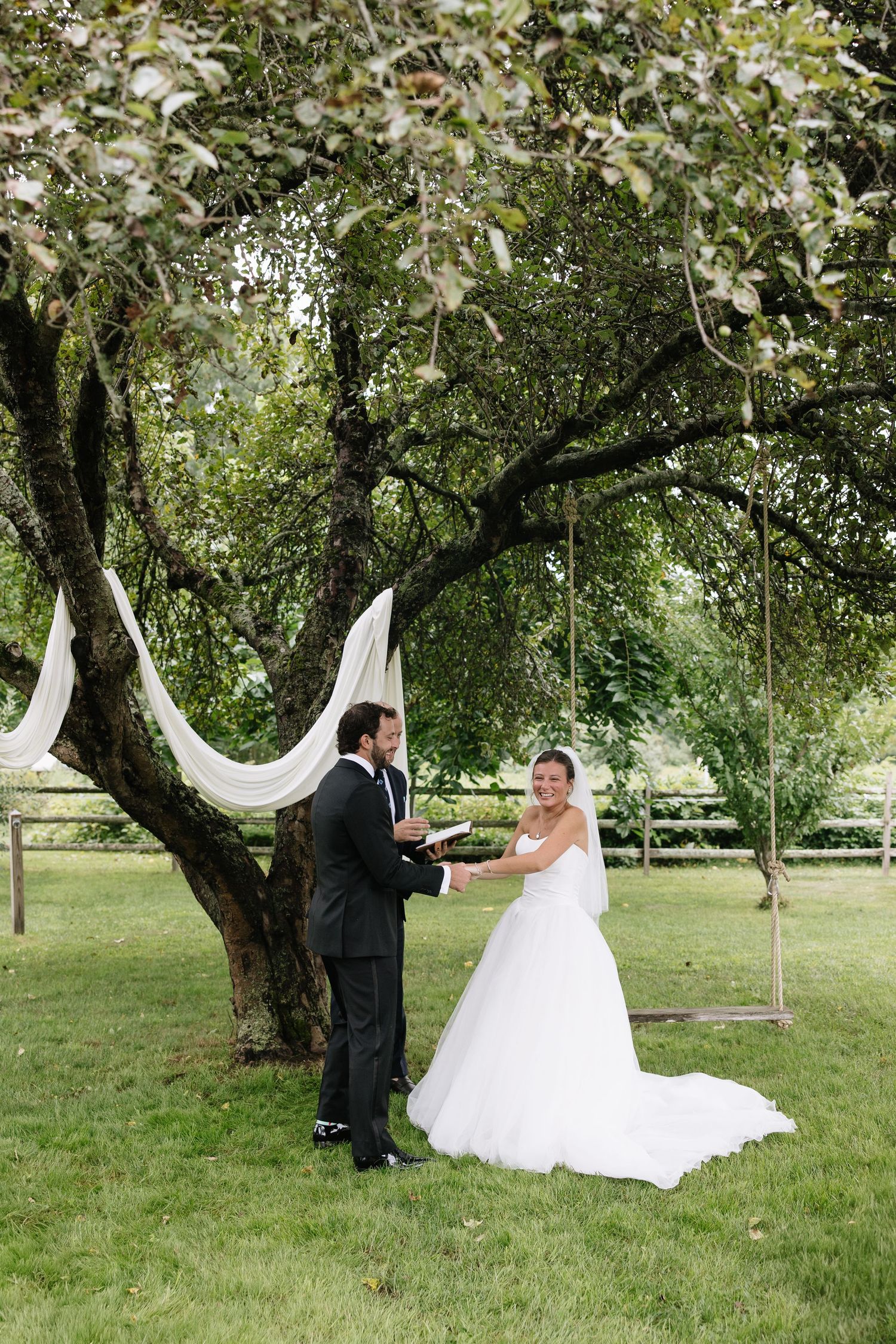 Bride and groom exchange vows under a decorated tree with white draping and a wooden swing.