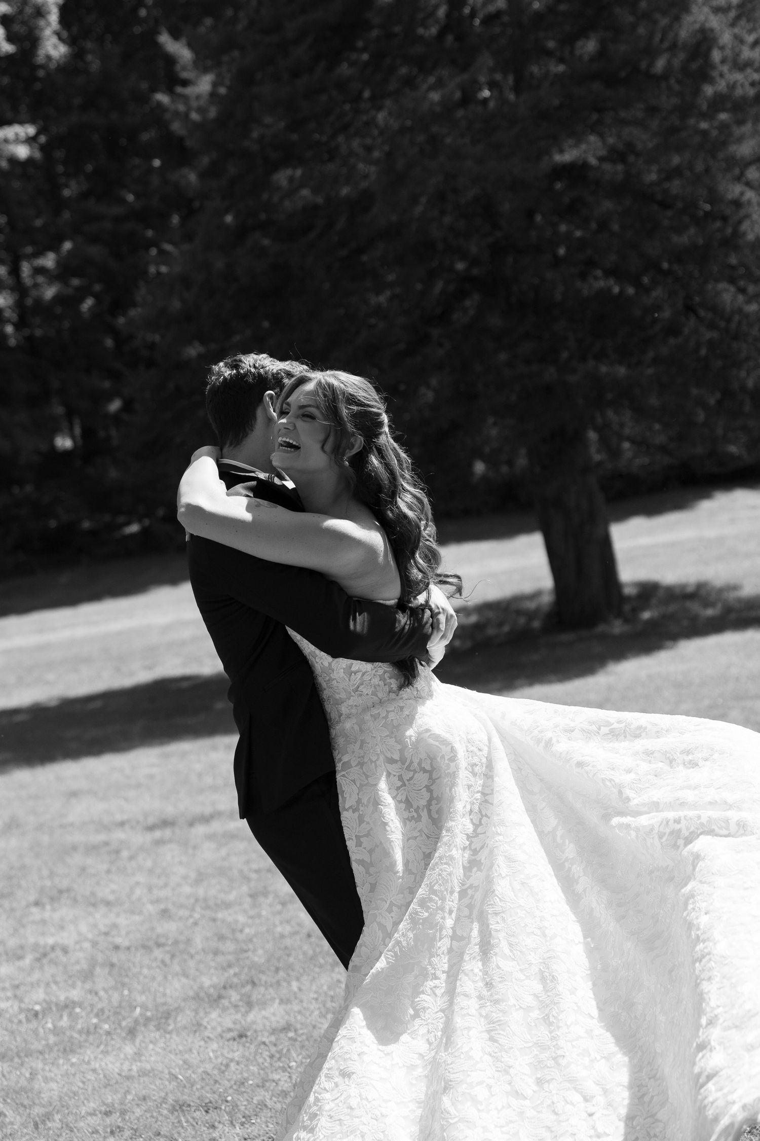 Black and white photo of a bride and groom embracing outdoors, her lace gown flowing as she smiles joyfully.