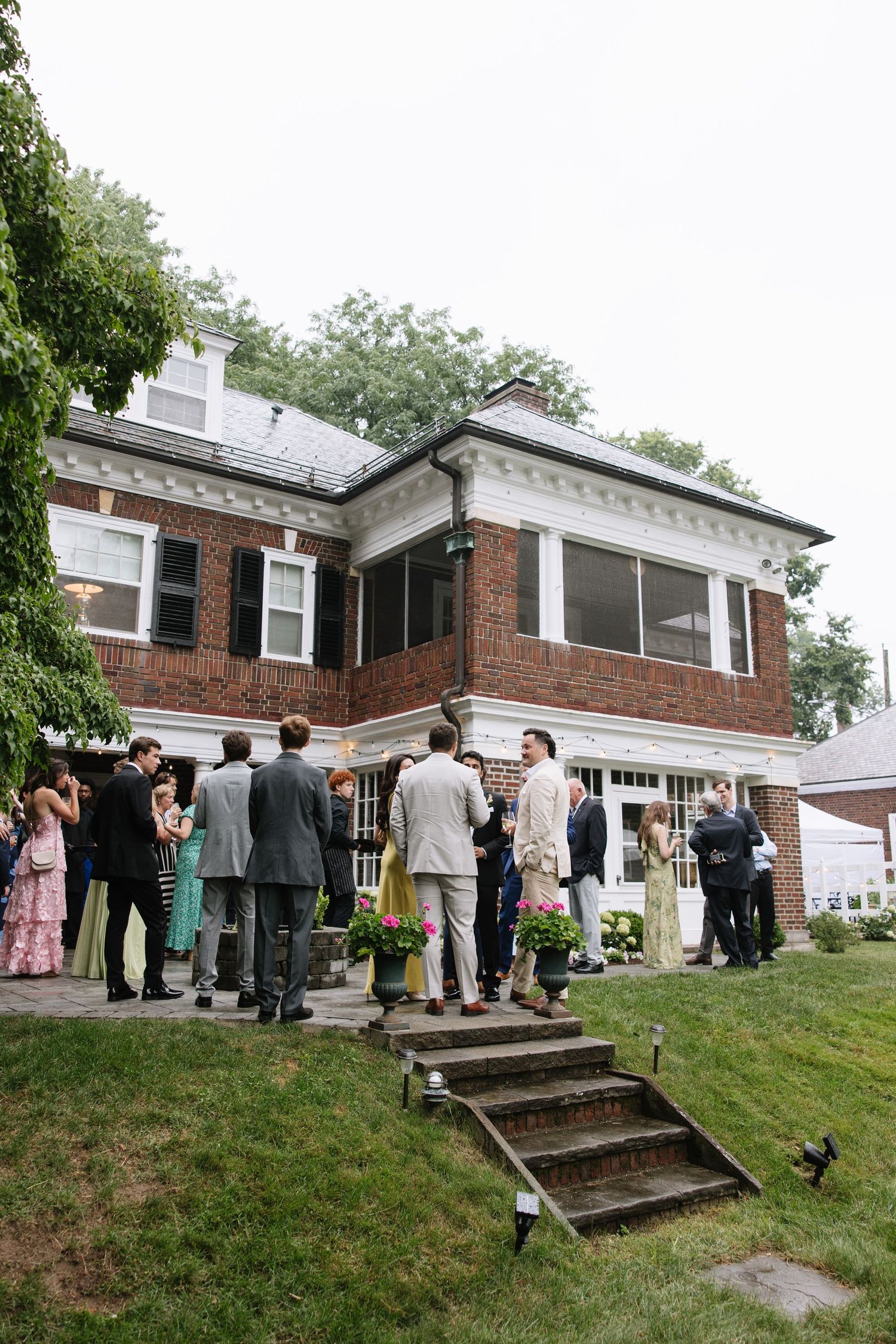 Wedding guests mingle outside a brick two-story mansion on a lush green lawn during a reception.