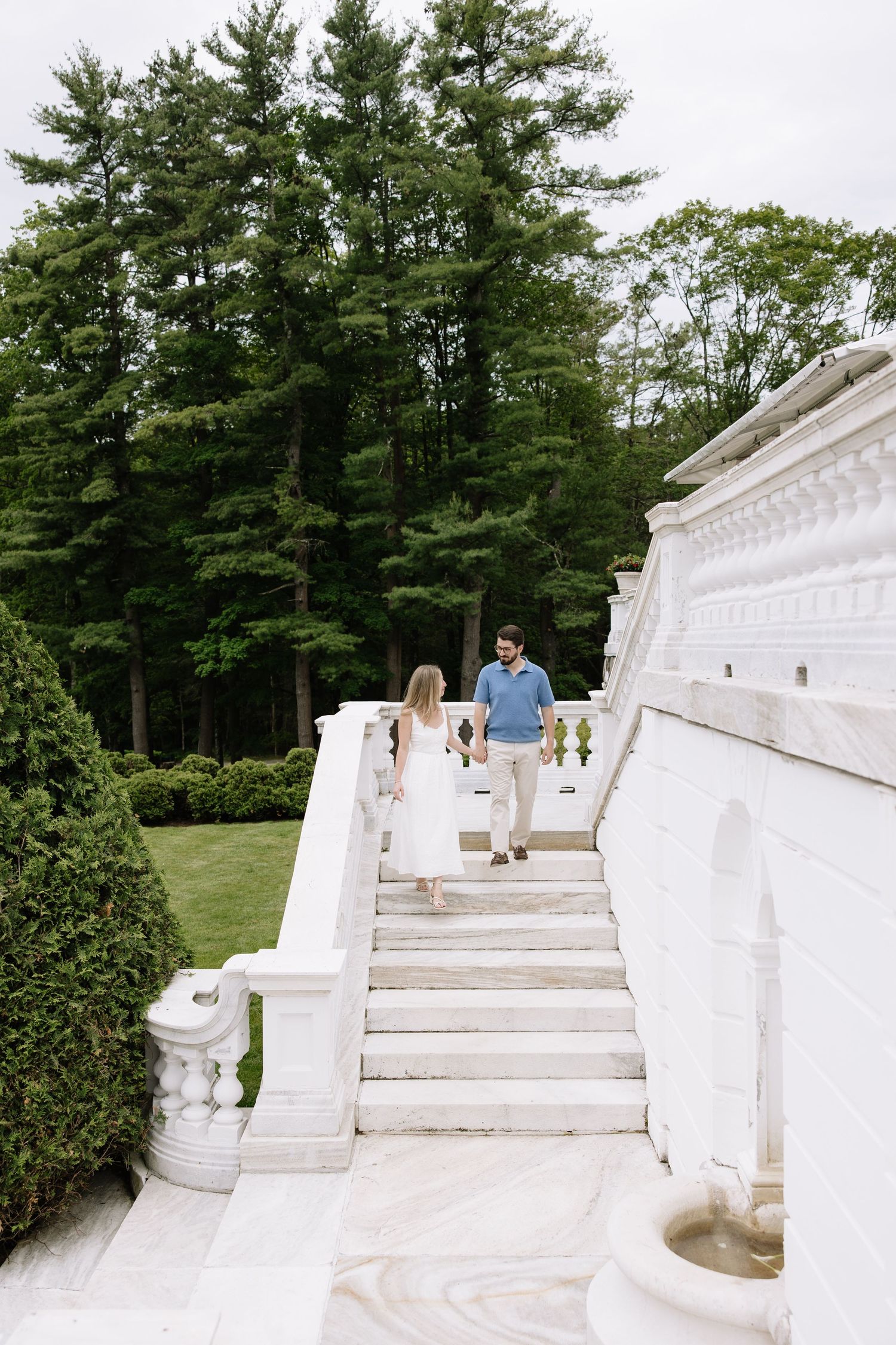Couple holding hands walking up elegant white marble stairs at a grand estate surrounded by tall pine trees.
