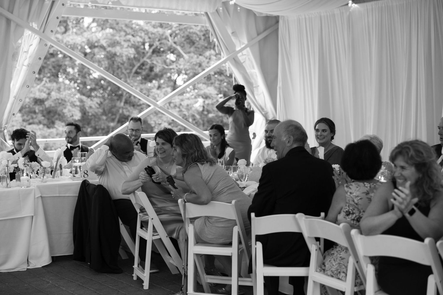 Black and white photo of wedding reception guests seated under elegant draped tent, socializing at decorated tables.