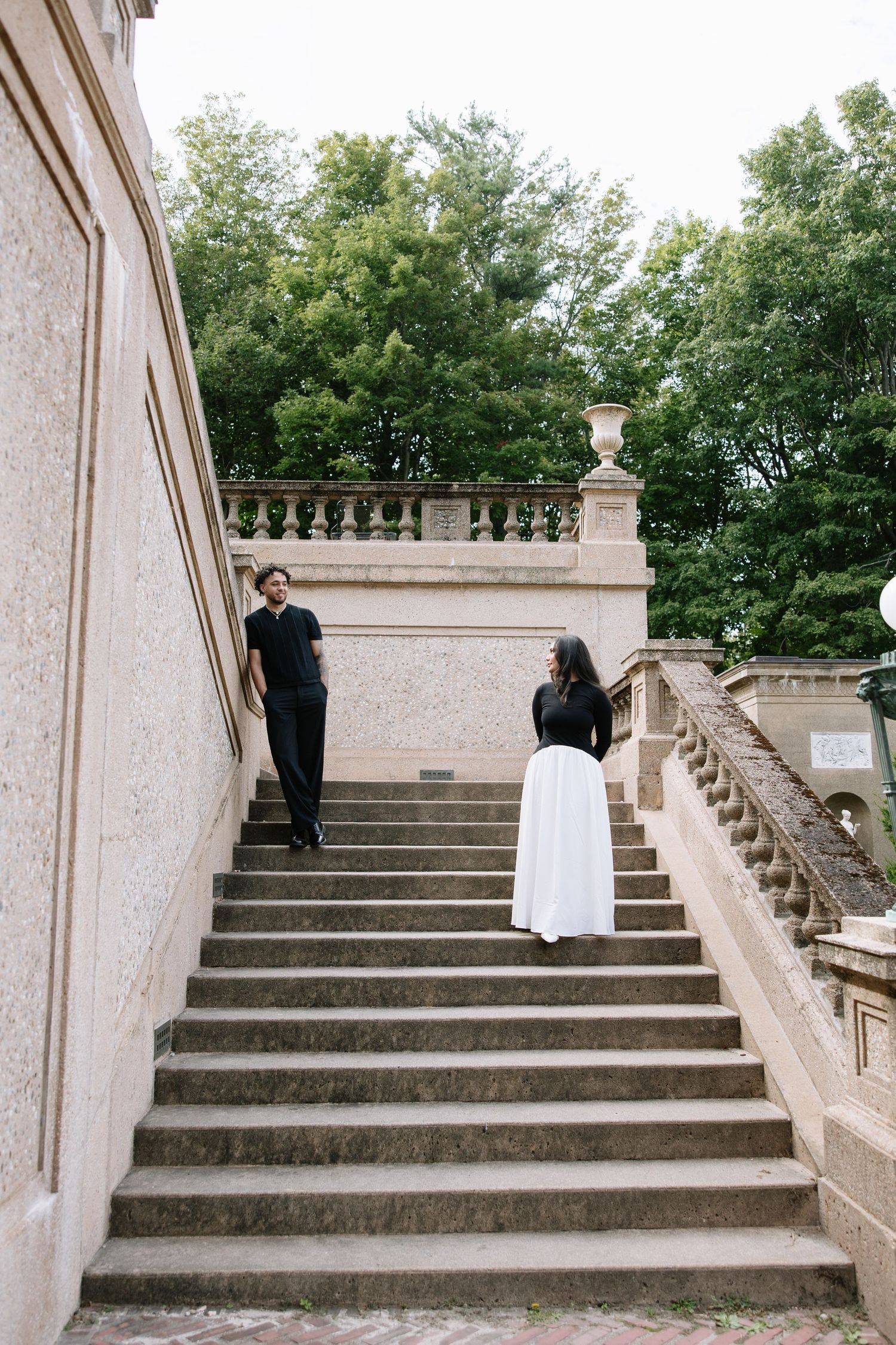 A couple in contrasting black and white outfits pose on an elegant stone staircase surrounded by greenery.