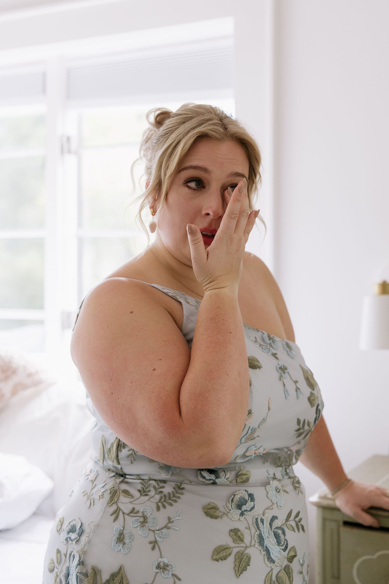 A woman in a floral dress shows surprise and emotion during a wedding preparation moment.