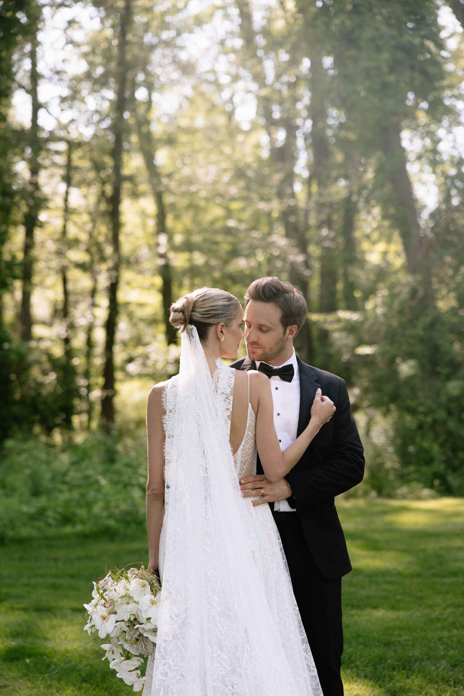 A romantic wedding moment captured in a sunlit forest setting with white floral bouquet details.