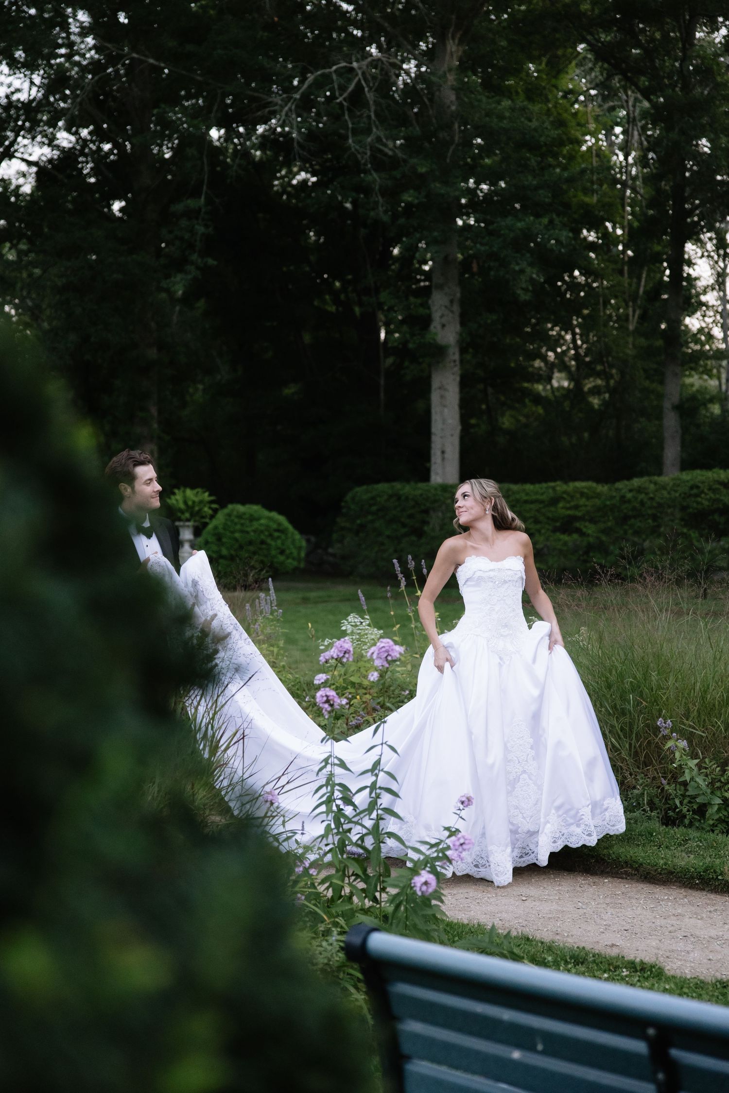 A bride in a flowing white dress twirls gracefully on a garden path surrounded by greenery.