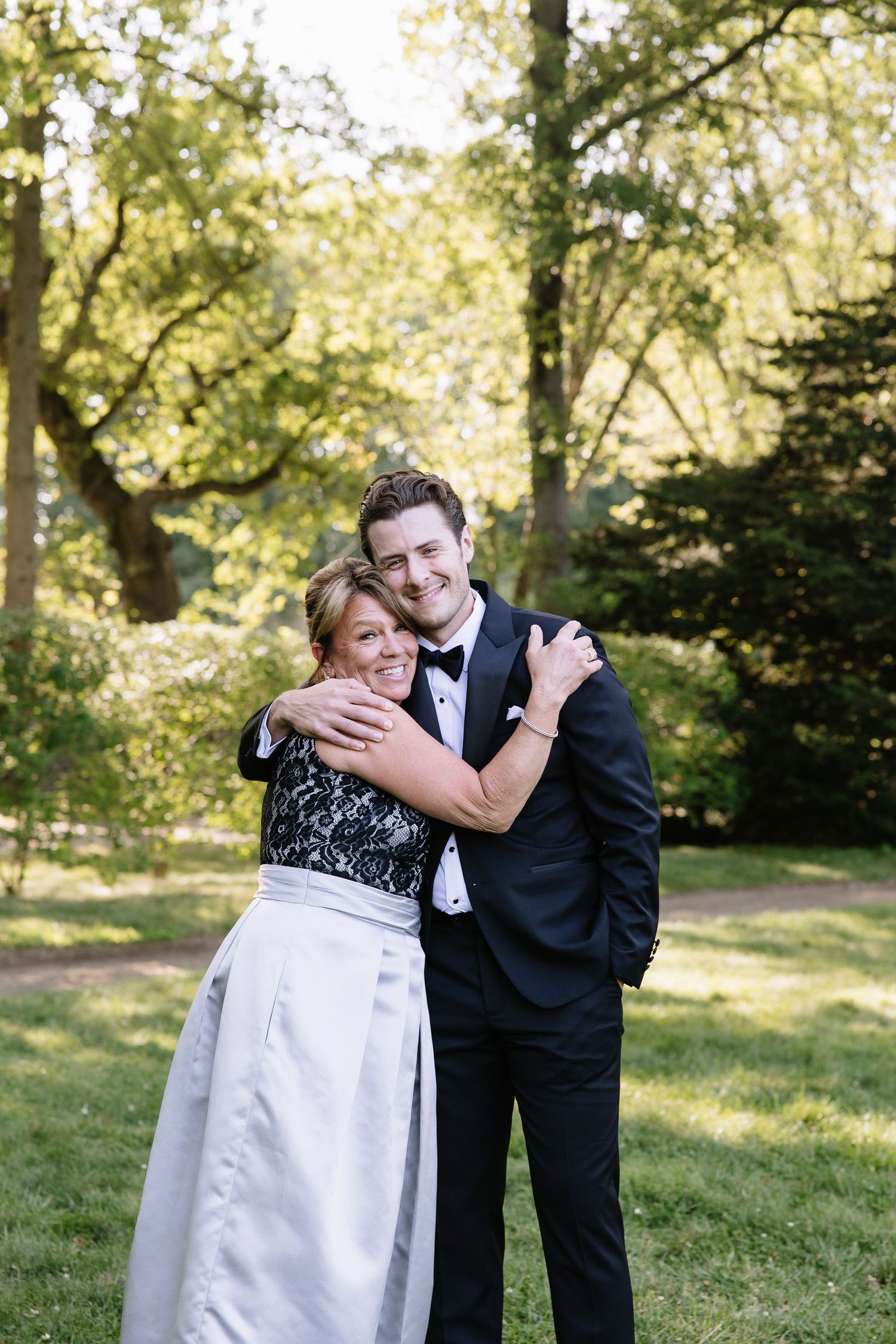 A couple shares a tender moment in formal attire at an outdoor wedding venue with lush greenery.