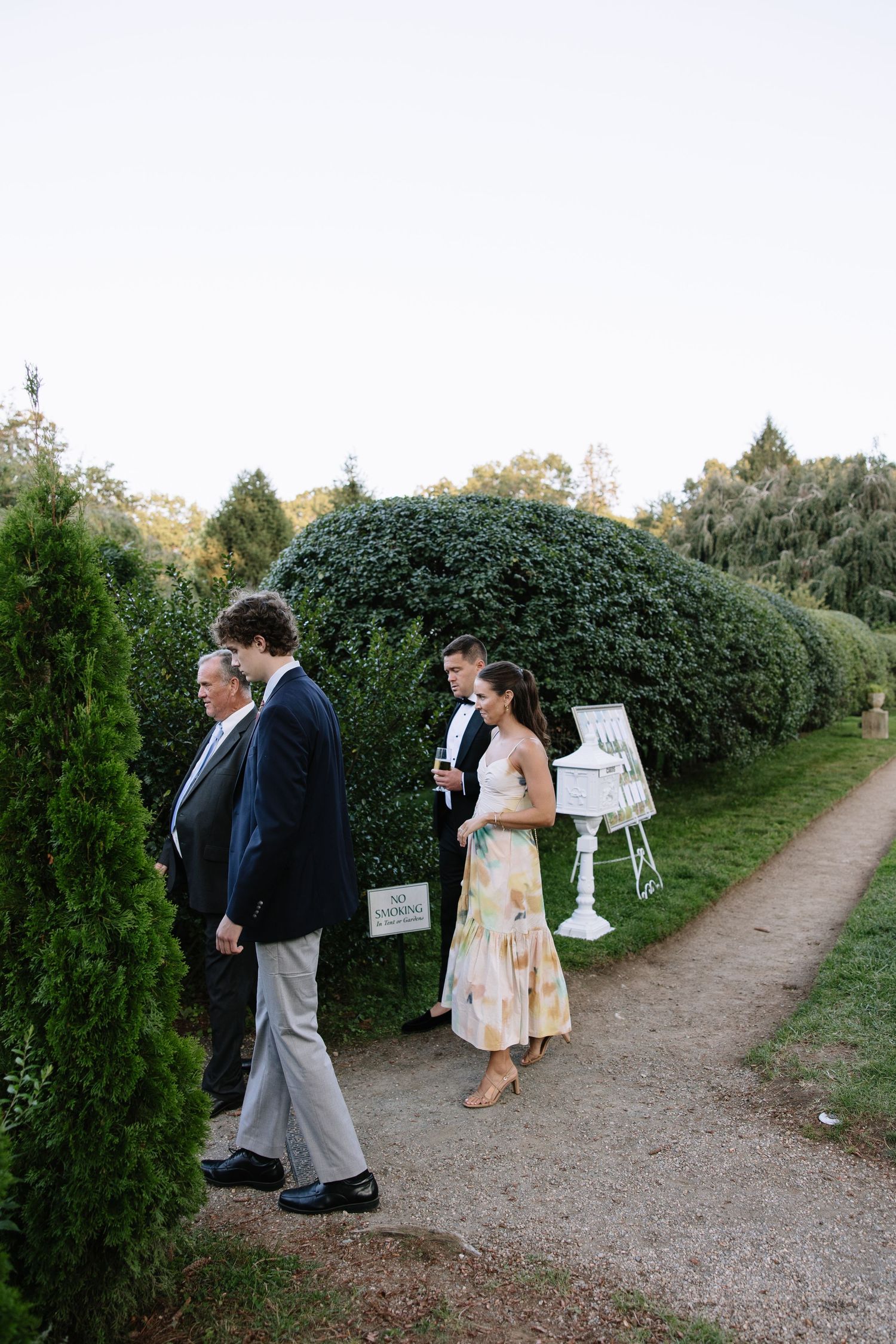 Wedding guests walk along a garden path lined with manicured hedges and trees.