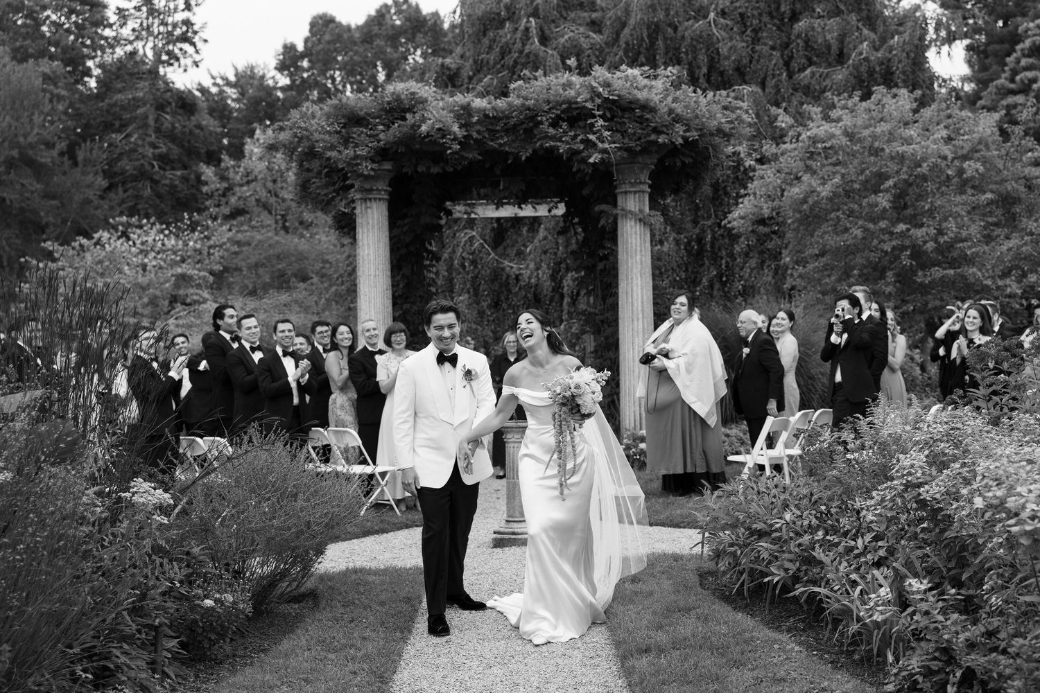 Newlywed couple in a white dress and tuxedo jacket walk down a garden aisle past smiling wedding guests beneath a vine-covered pergola.