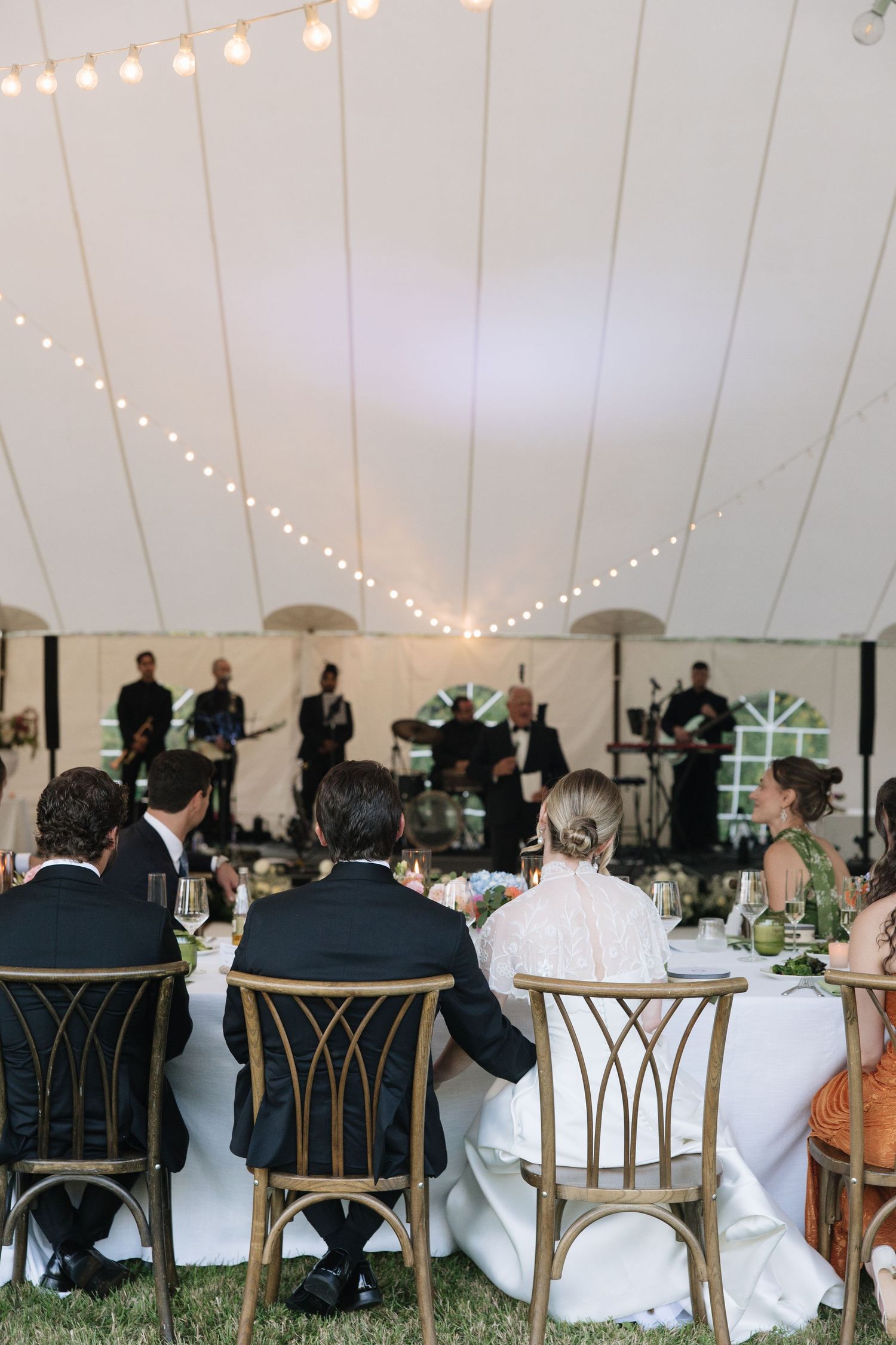 Guests seated at wedding reception tables under a white tent with string lights and a live band performing on stage.