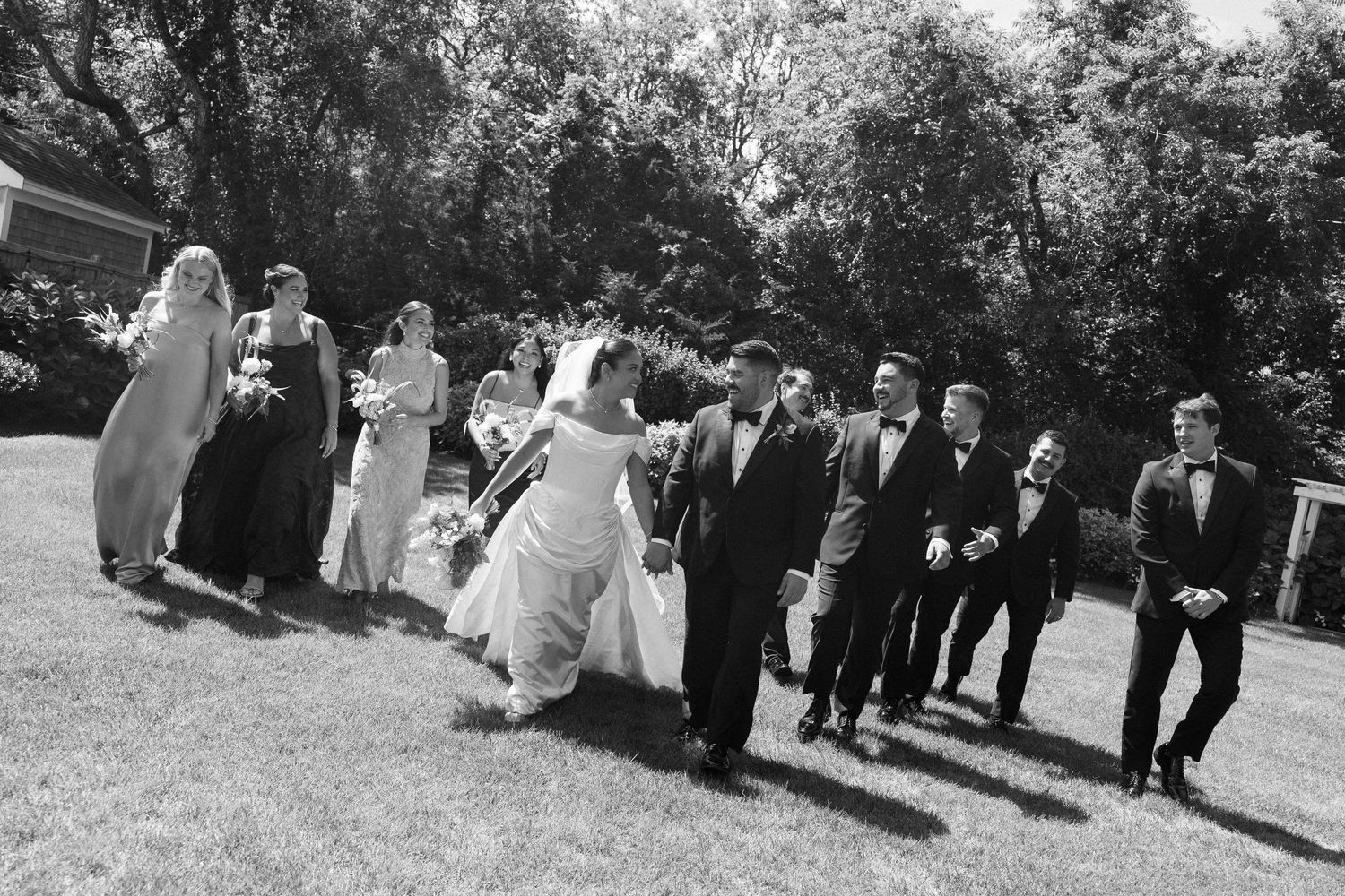 Black and white photo of a wedding party walking across a green lawn, bride in white gown with groom and attendants.