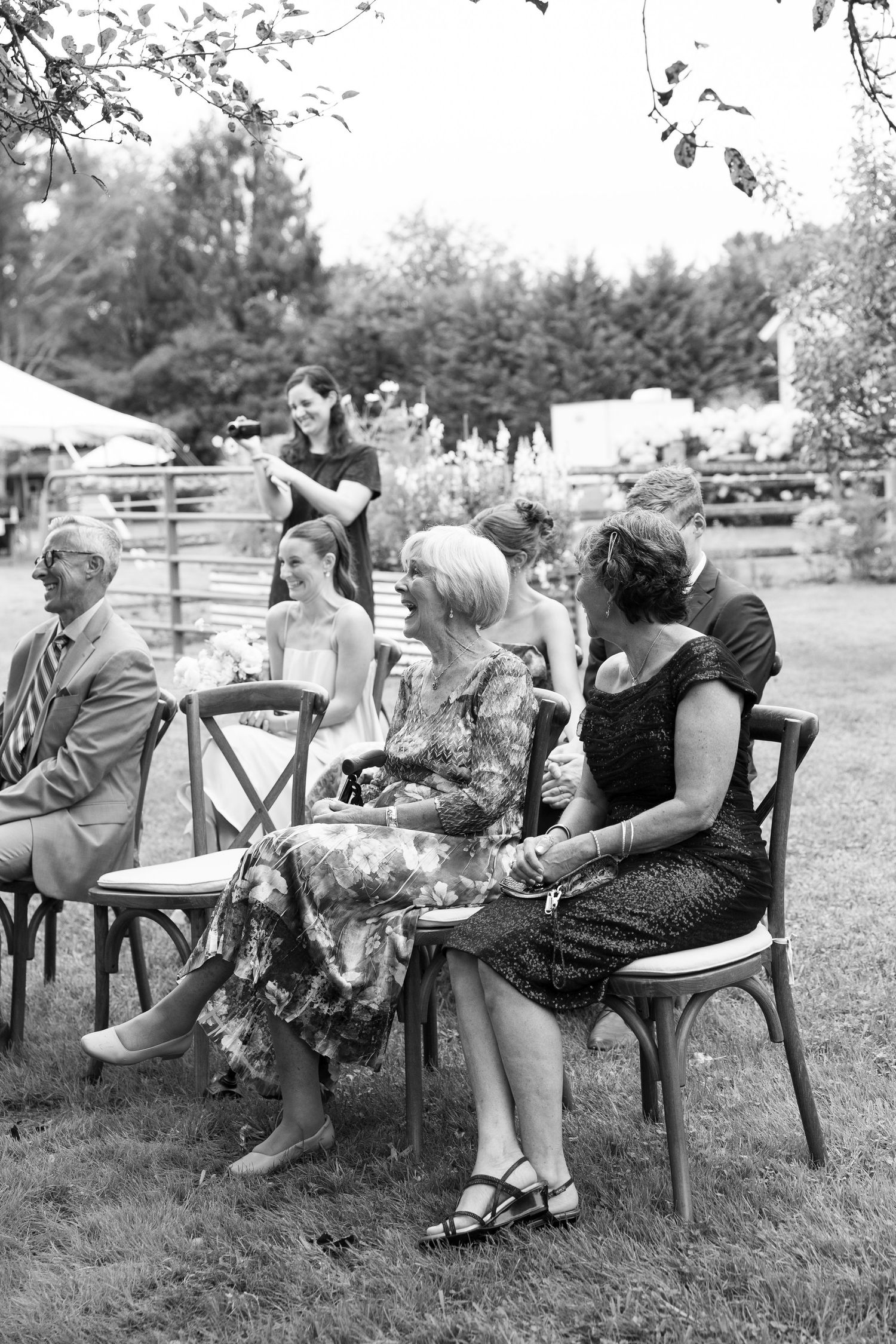 Guests seated outdoors on wooden chairs at an elegant garden wedding ceremony in black and white.
