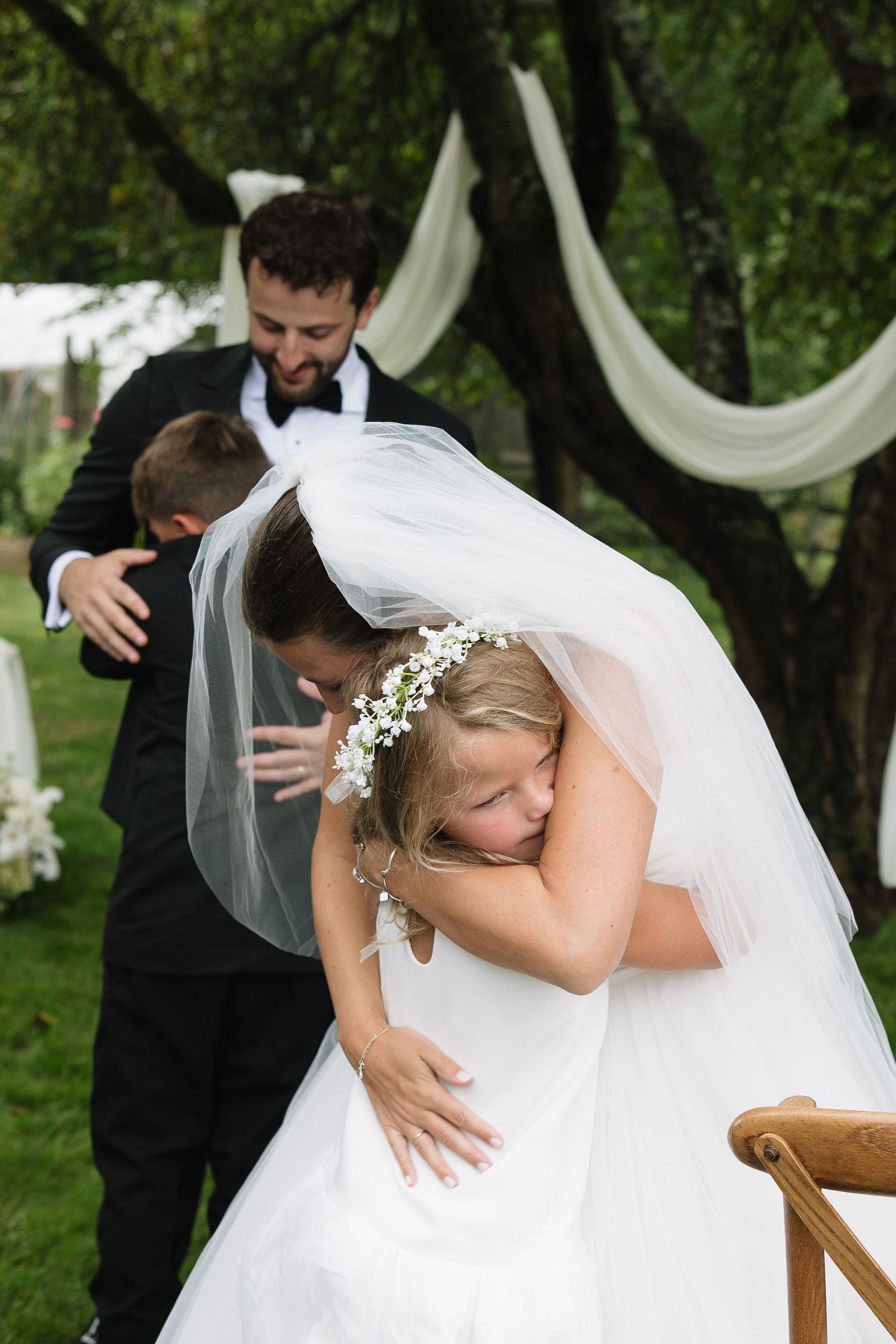 Bride hugging a child during outdoor wedding ceremony while groom smiles in background.