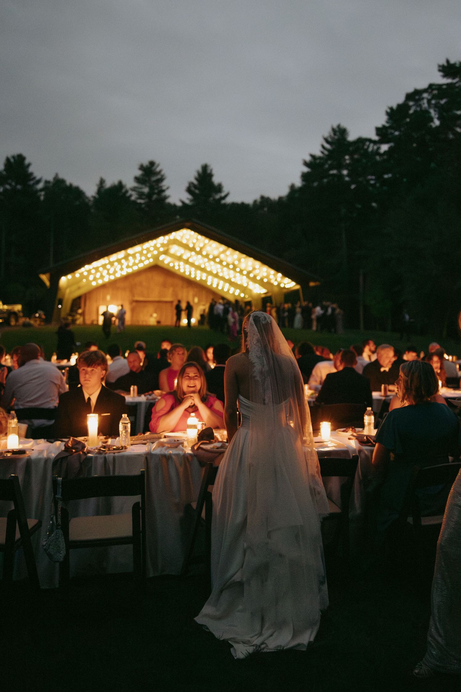 Bride in white gown and veil stands among candlelit outdoor wedding reception tables at dusk near illuminated pavilion.