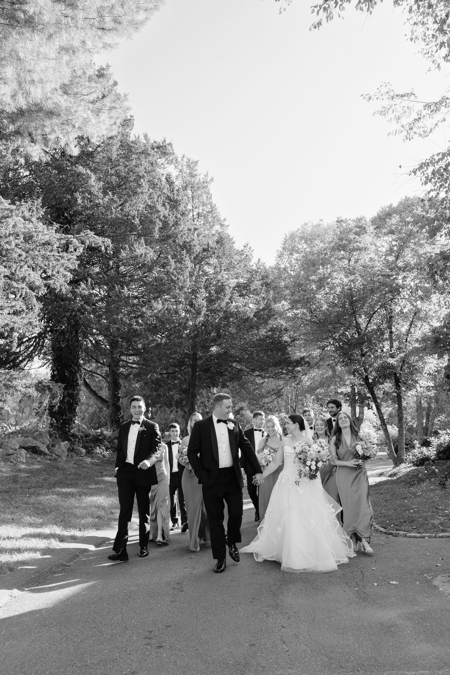 Black and white photo of a wedding party walking outdoors, bride in gown holding bouquet, groom in tuxedo, among tall trees.