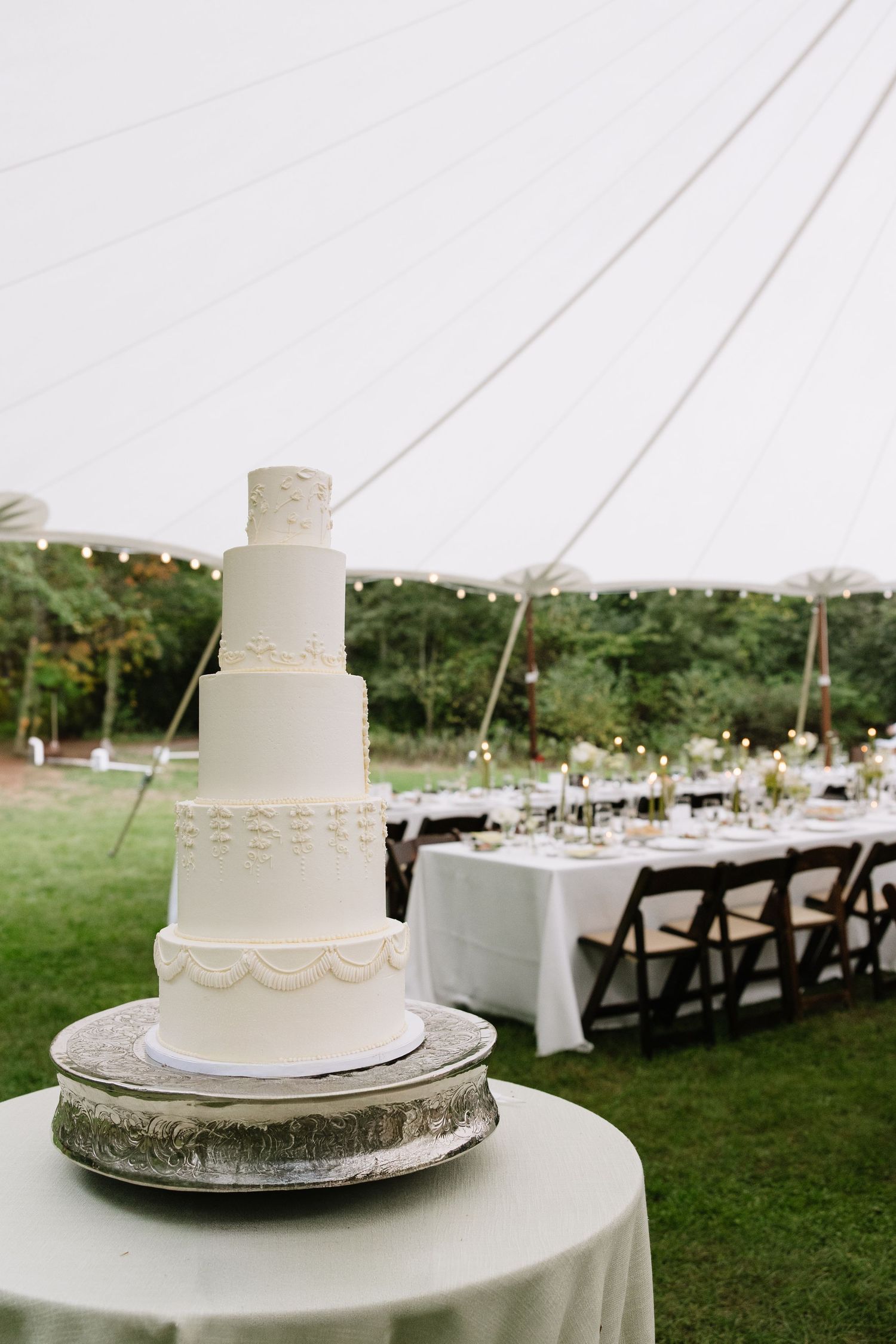 Elegant five-tier white wedding cake on silver stand under a white sailcloth tent with string lights and reception tables.