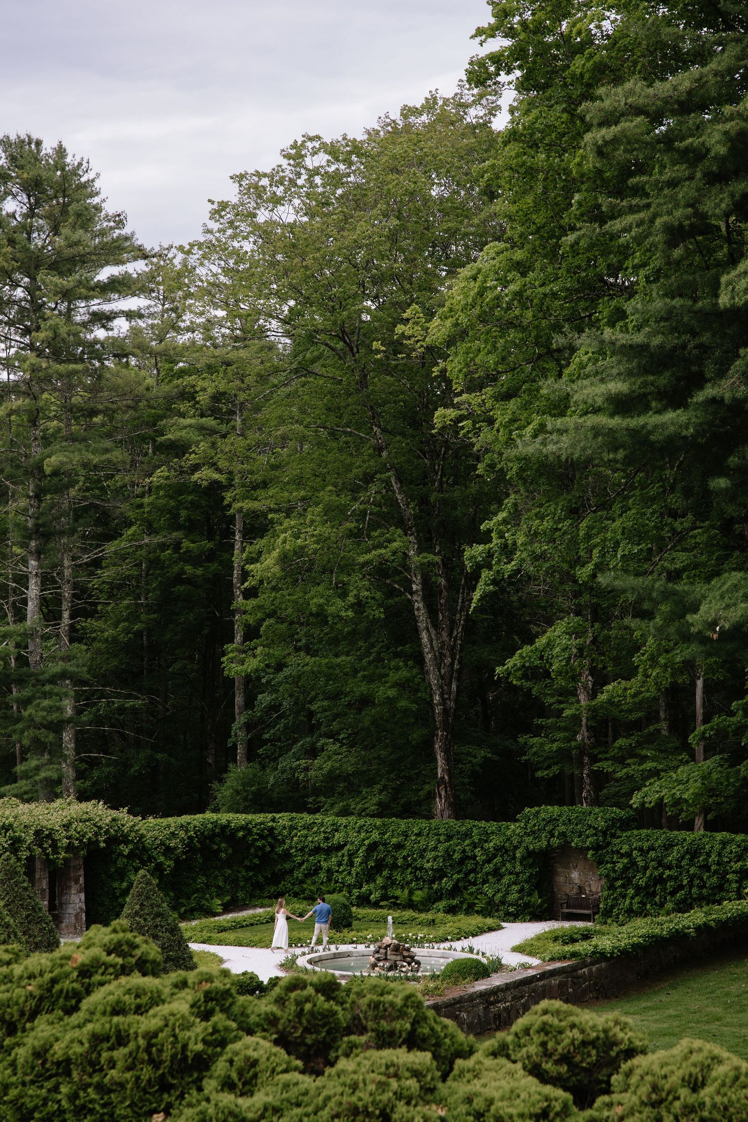 Couple in formal garden with manicured hedges and topiary surrounded by dense green forest.