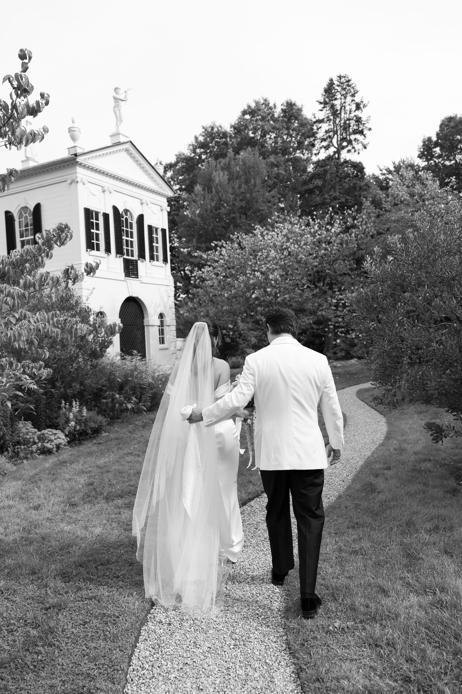 Black and white photo of bride and groom walking hand in hand toward elegant white estate surrounded by lush gardens.