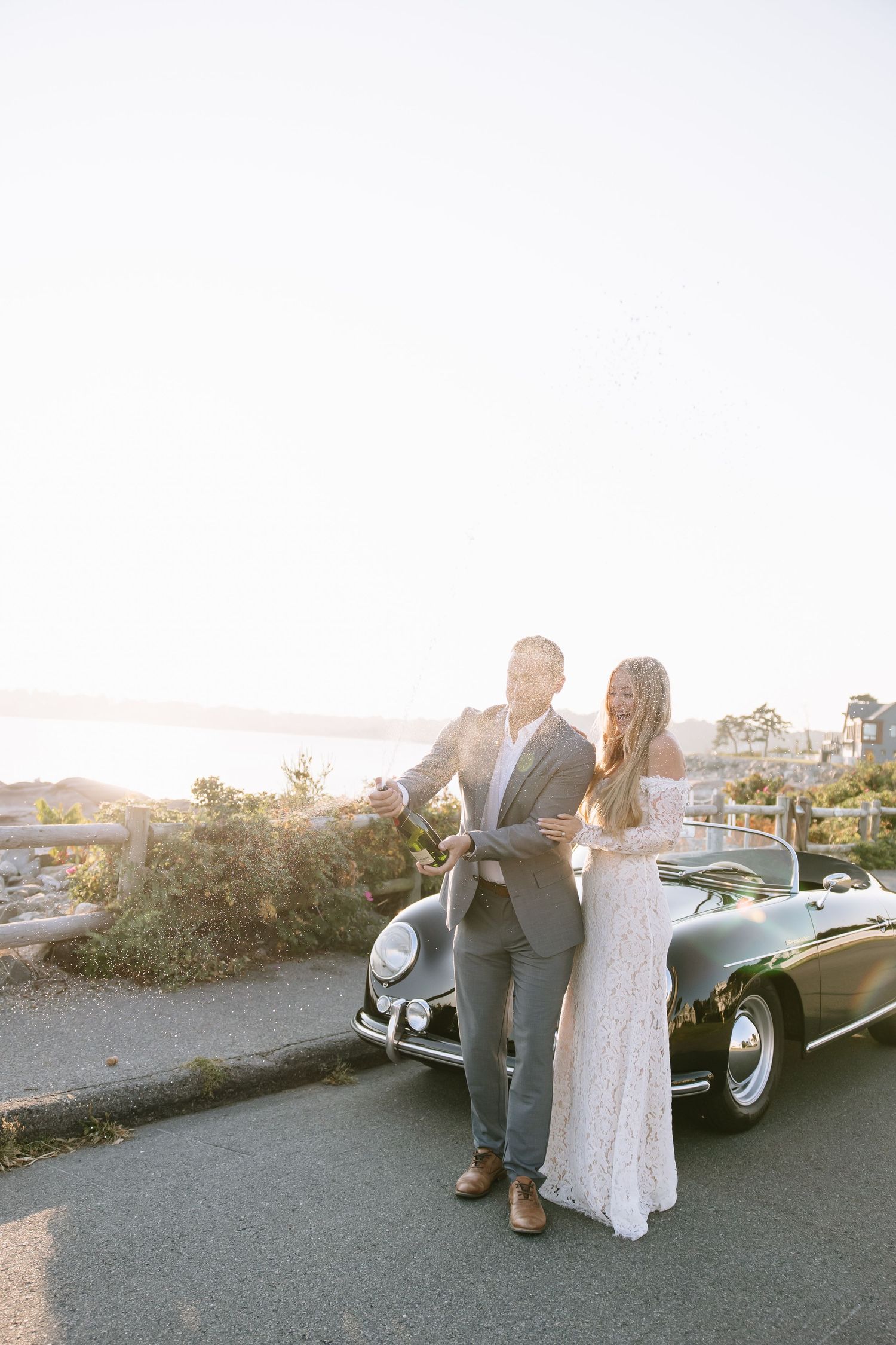 Bride and groom standing by a vintage convertible car at sunset with ocean views in the background.