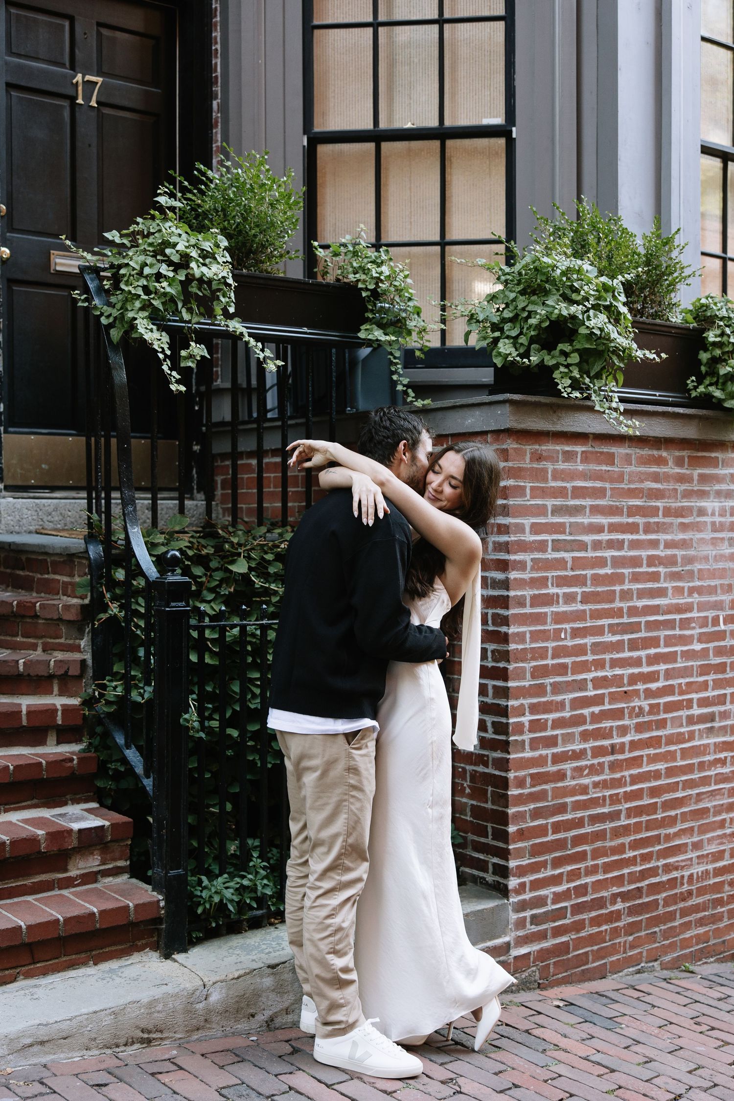 Couple embracing against brick townhouse with window planters and black railings.
