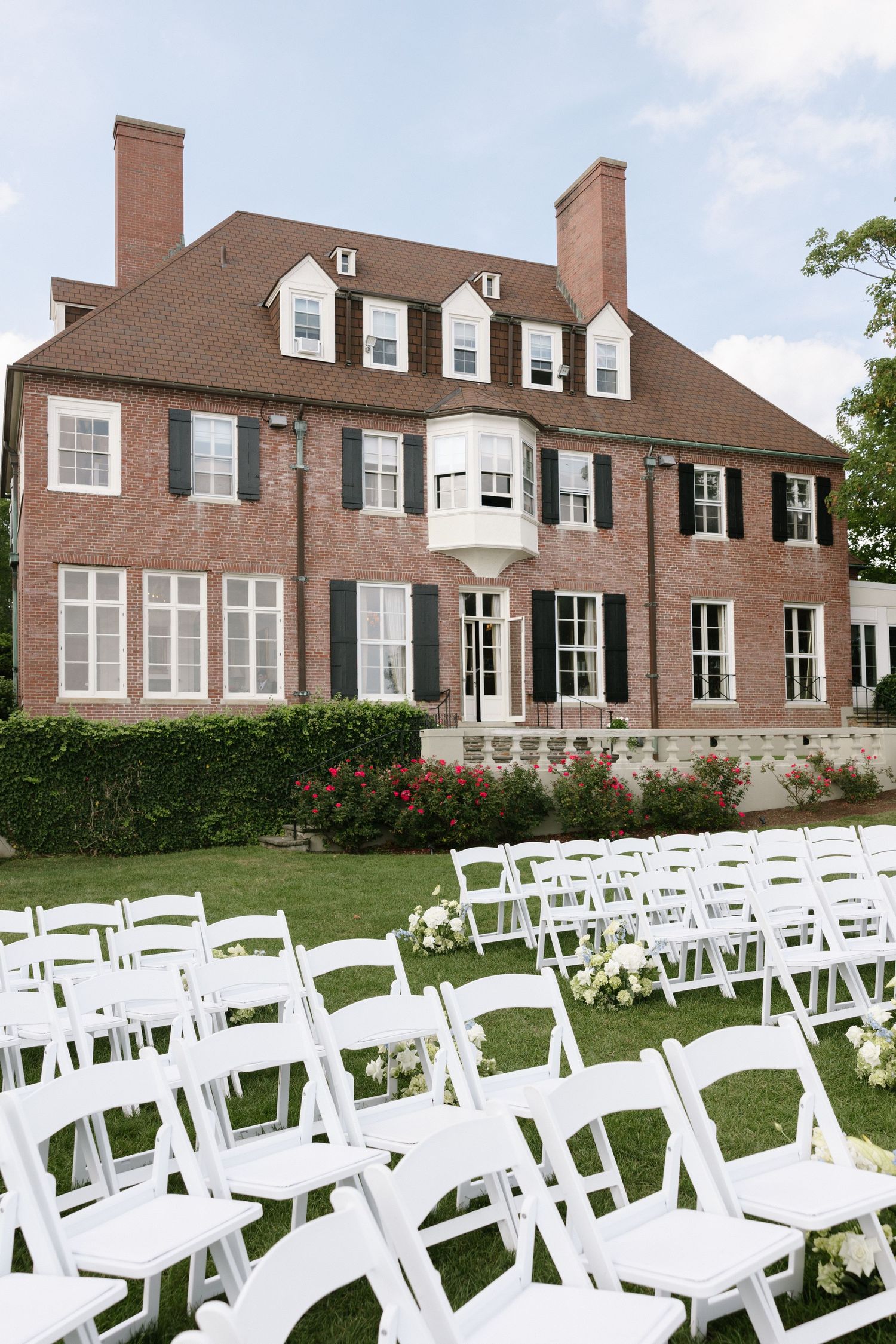 Rows of white folding chairs with flower arrangements face a red brick manor house with dormer windows and black shutters.