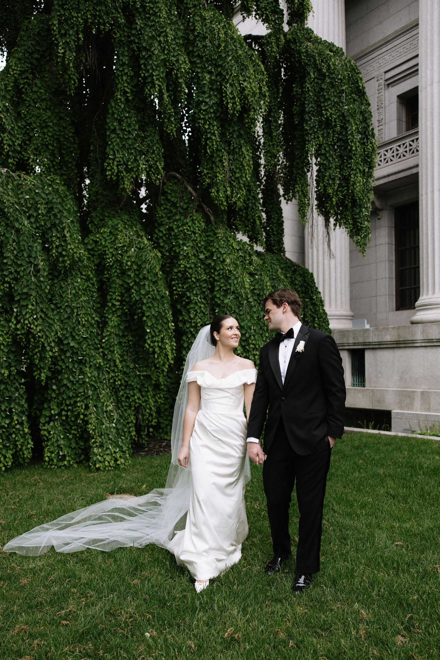 Bride in white gown and veil walks with groom in black tuxedo on green lawn before ivy-covered building.