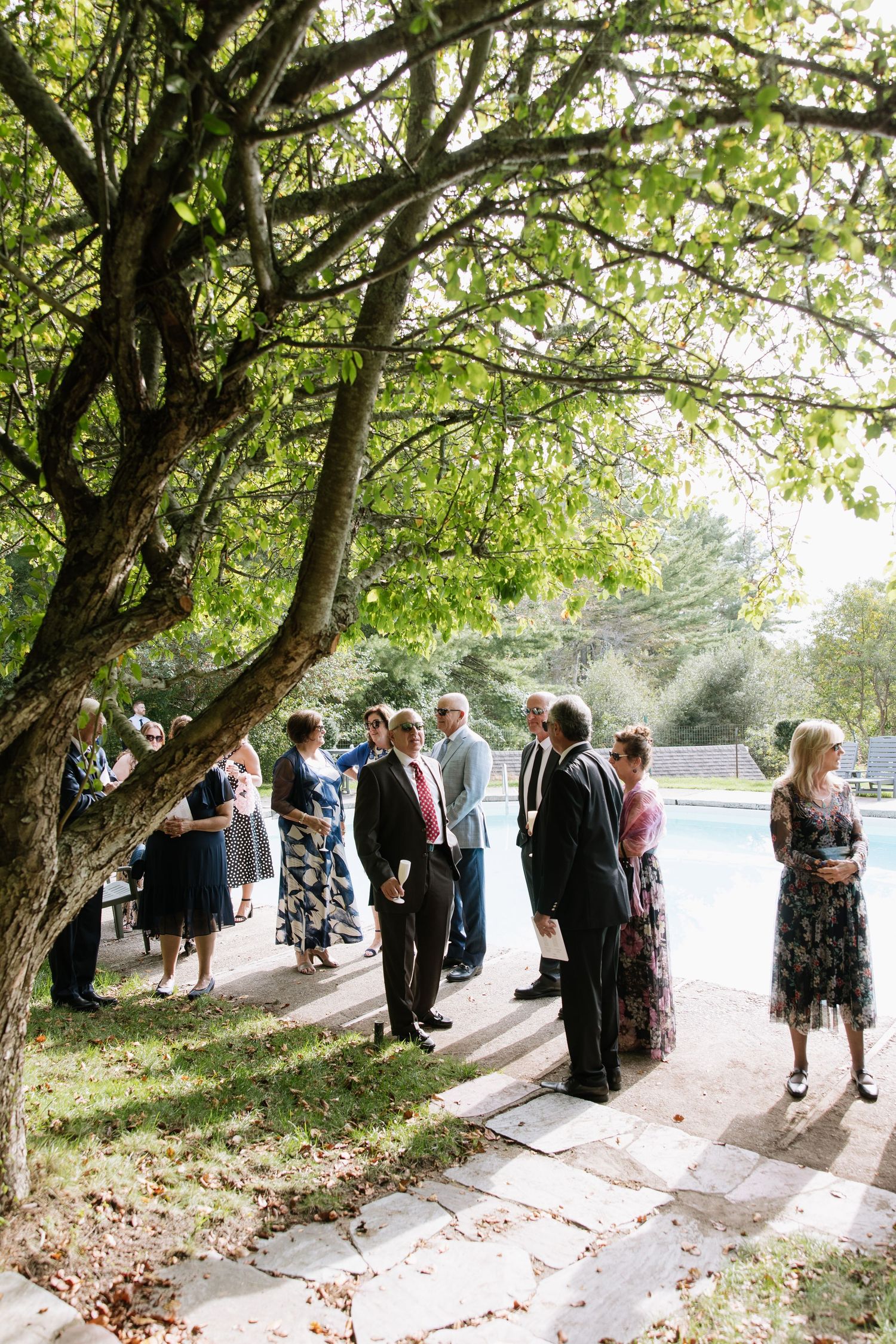 Elegantly dressed wedding guests mingle outdoors near a pool, surrounded by lush trees on a stone pathway.