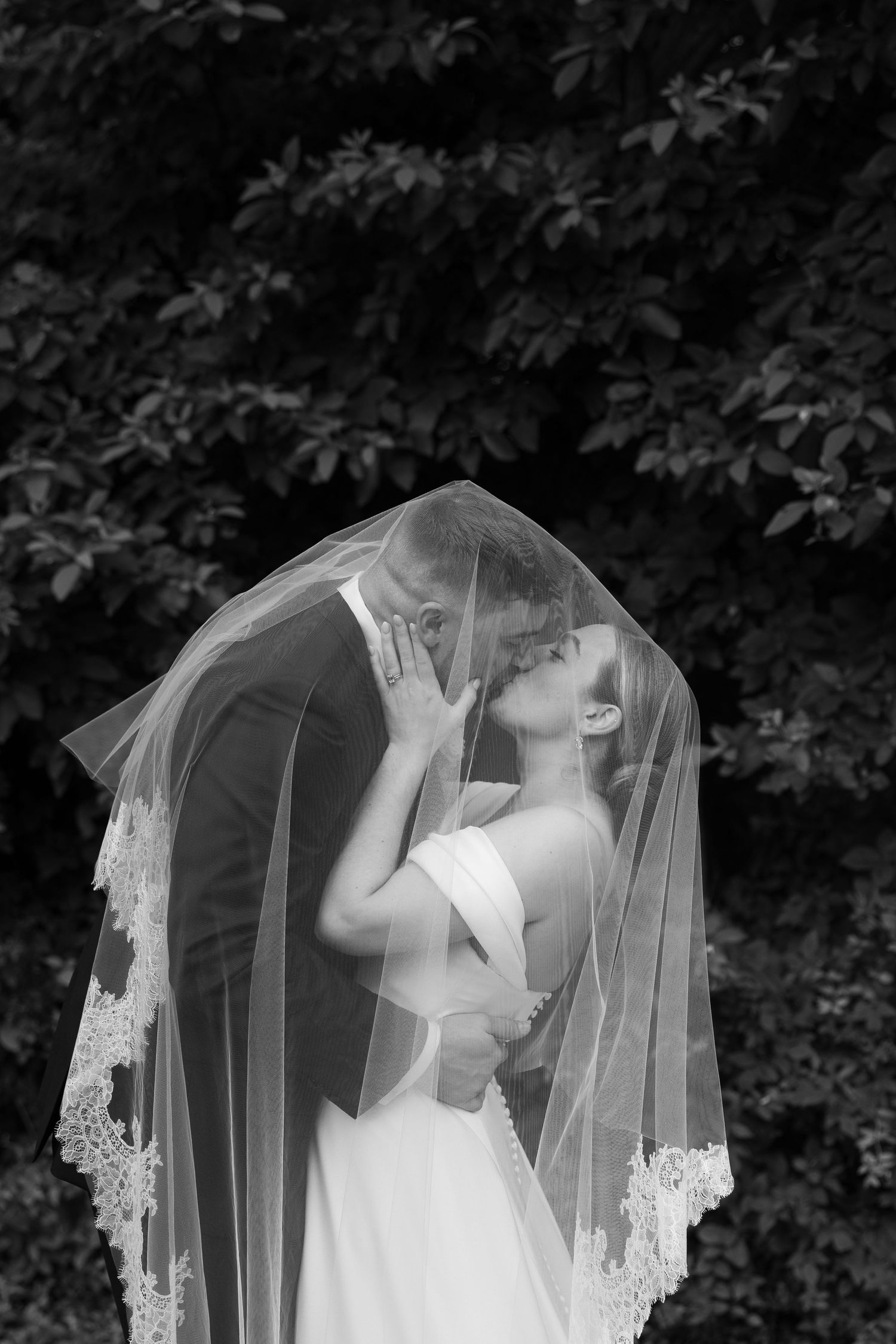 Black and white photo of bride and groom kissing under a lace veil outdoors.