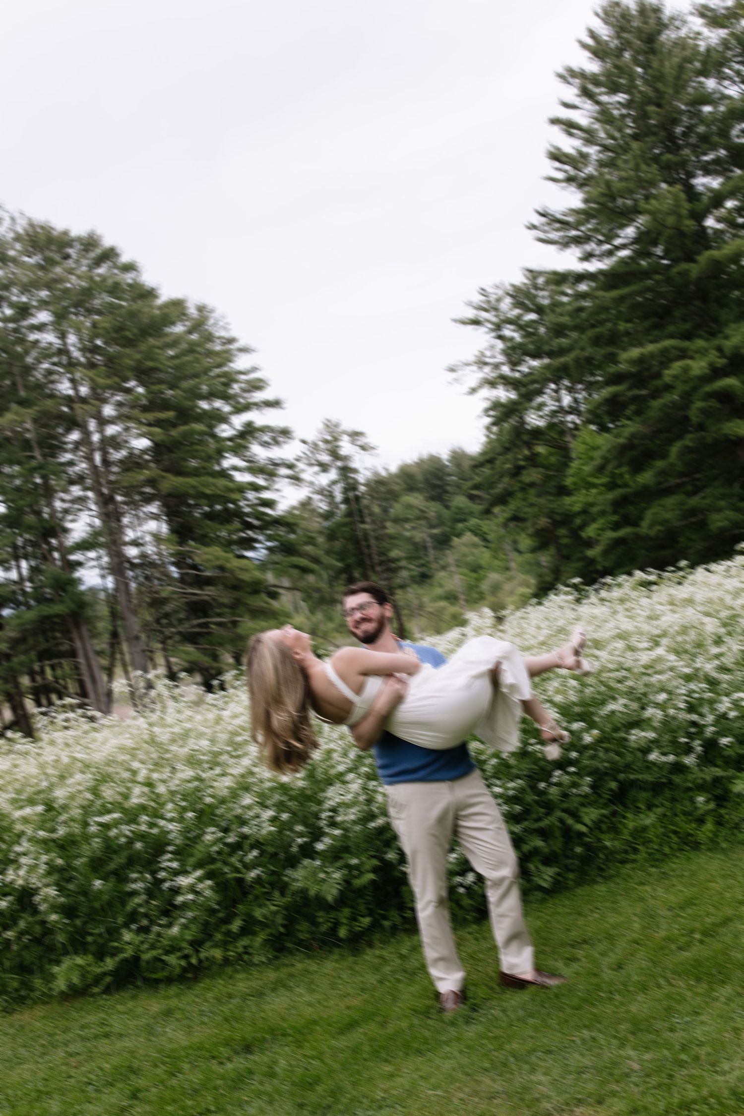 Man lifting and spinning woman in lush garden with white wildflowers and tall evergreen trees.