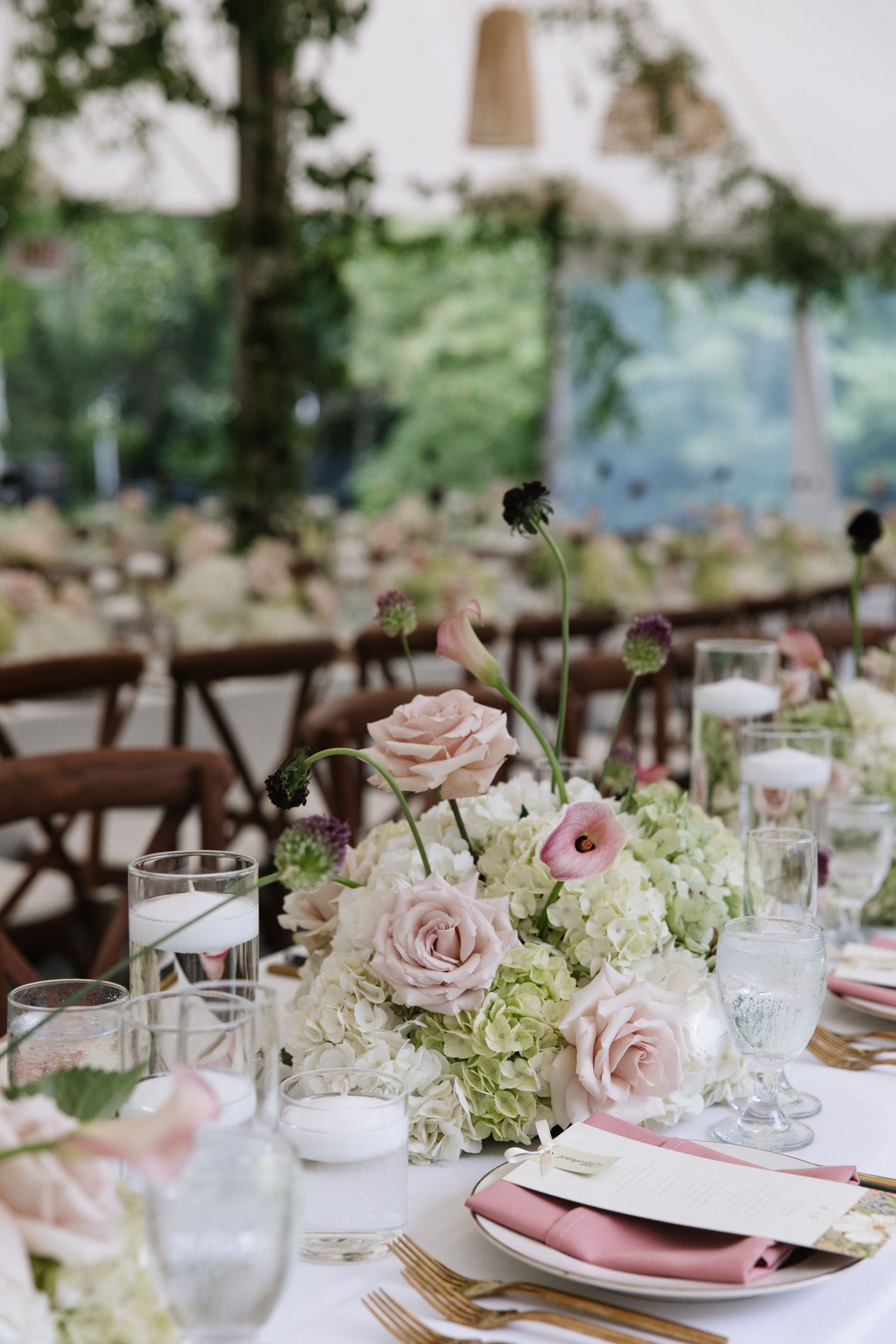 Elegant wedding reception table with blush roses, white hydrangeas, candles, pink napkins, and gold flatware under a tent.