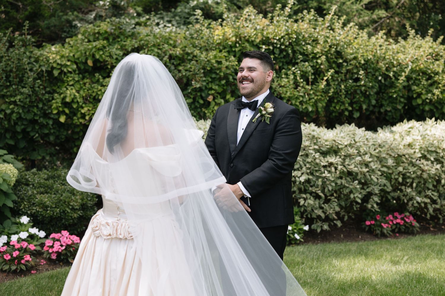 Groom in black tuxedo smiles at bride in white gown and veil during outdoor first look in garden.