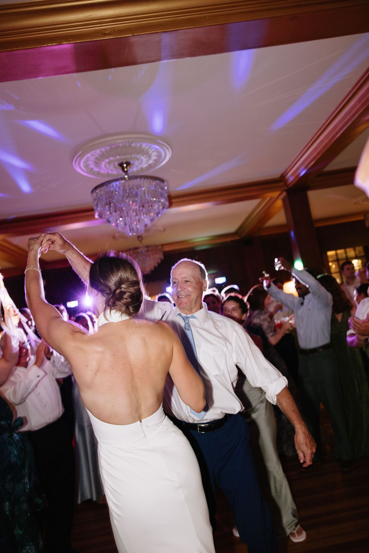 Bride in backless gown dances with older man in white shirt at elegant wedding reception.