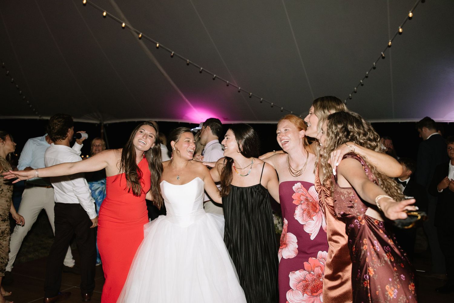 Bride in white ballgown dances joyfully with bridesmaids and friends under tent at wedding reception.